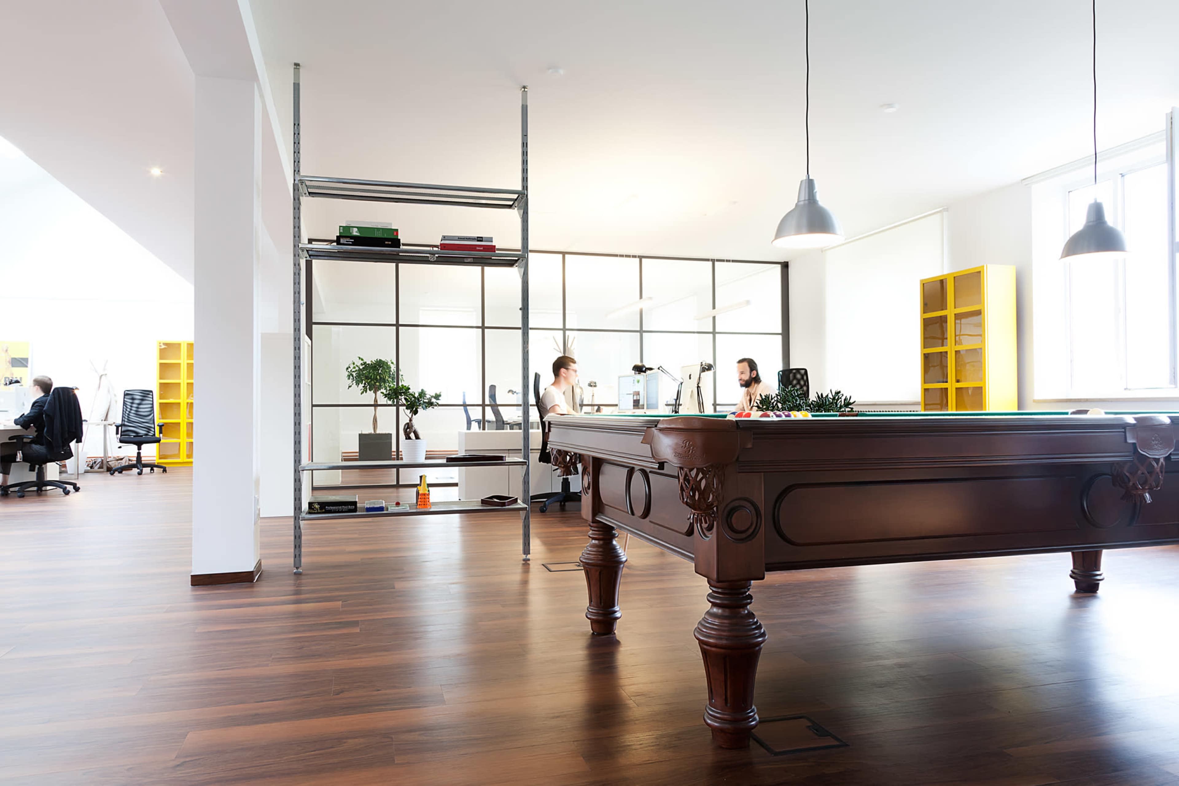 Two women are seated at a table in a modern office, with a billiards table in the foreground and shelves lined with books and plants in the background.