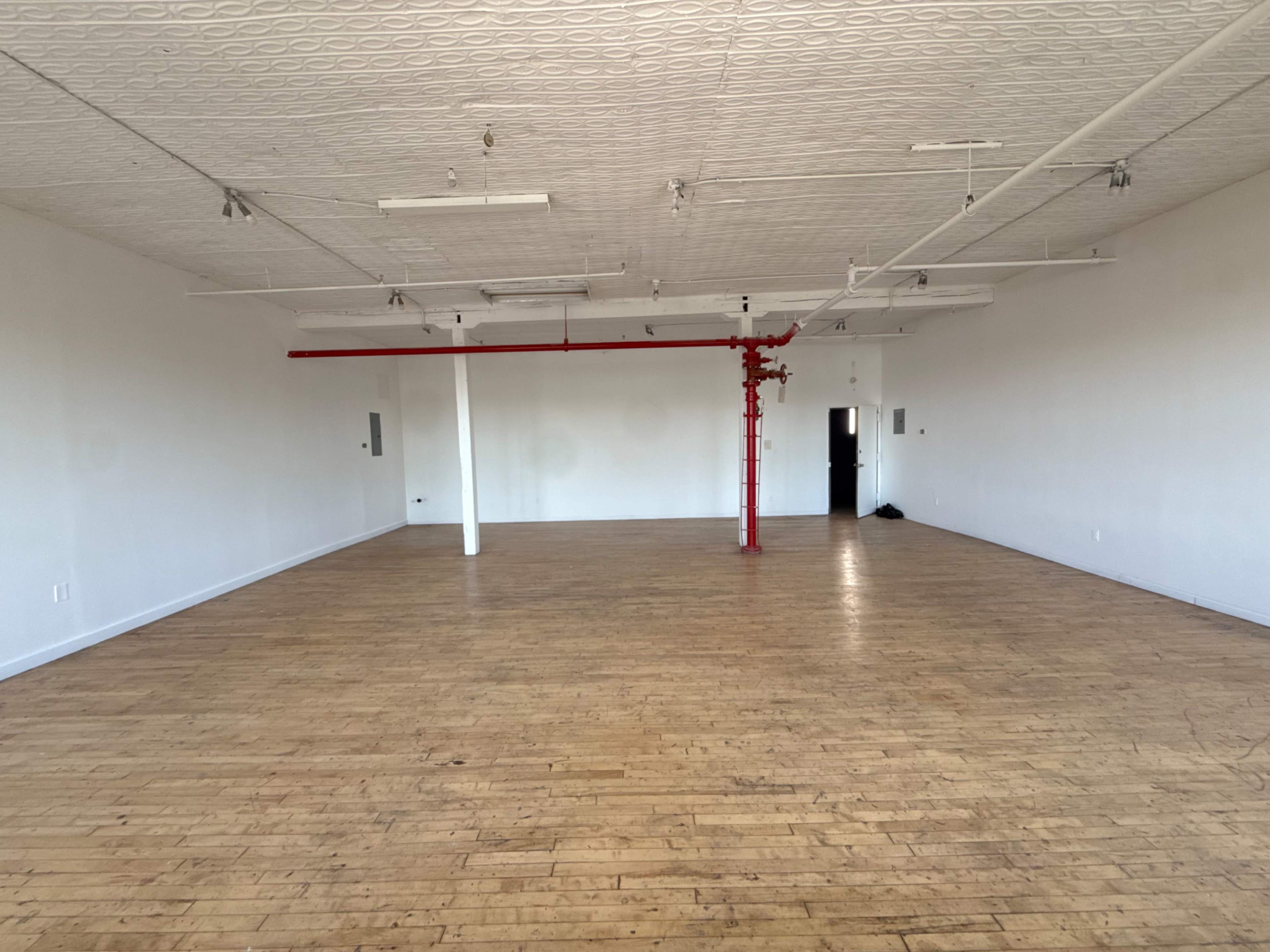The image shows a spacious, empty room with wooden flooring and minimal furnishings, featuring a red pipe along the ceiling.