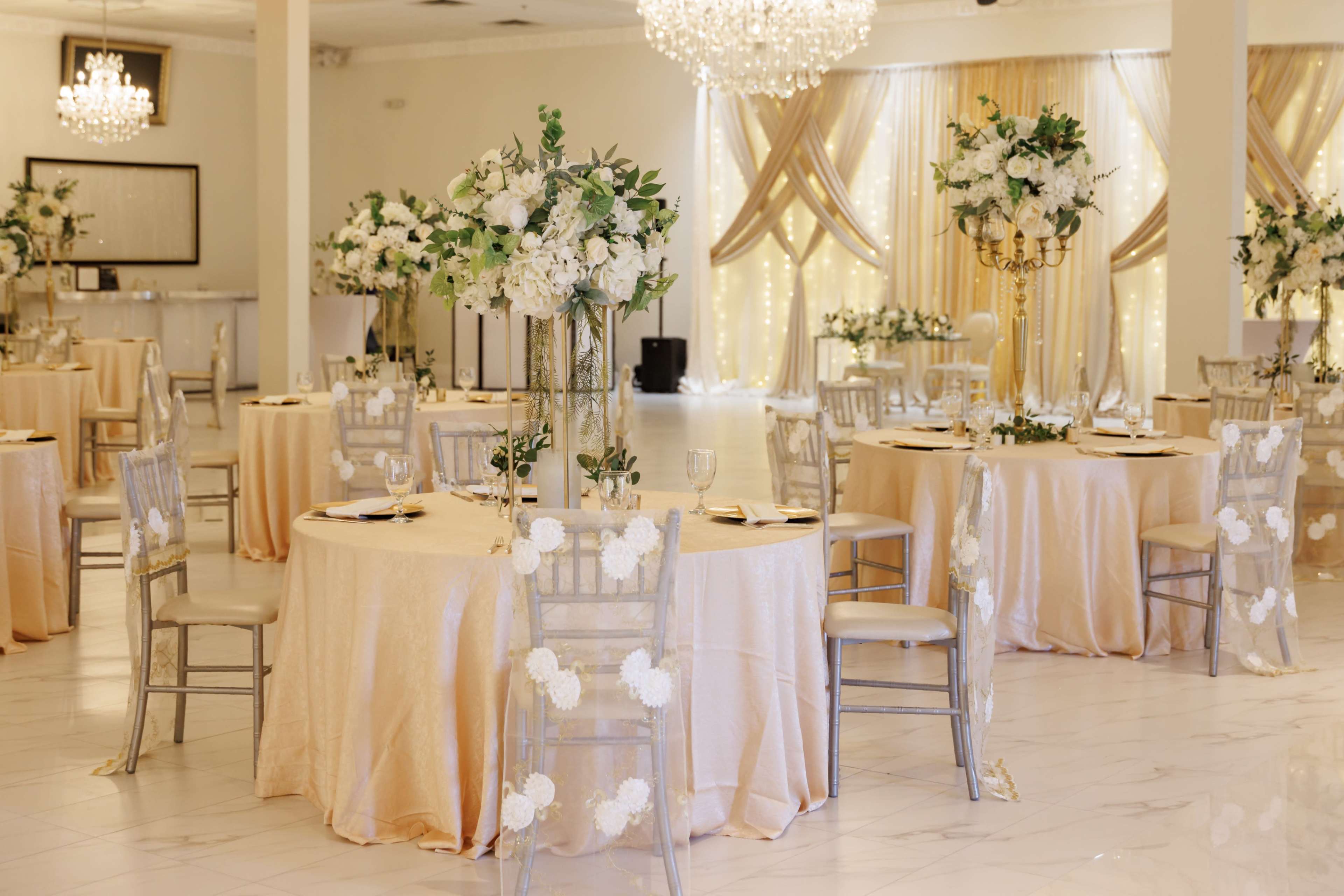 A banquet hall features elegantly set tables with gold tablecloths and floral centerpieces, complemented by chandeliers and decorative drapes.