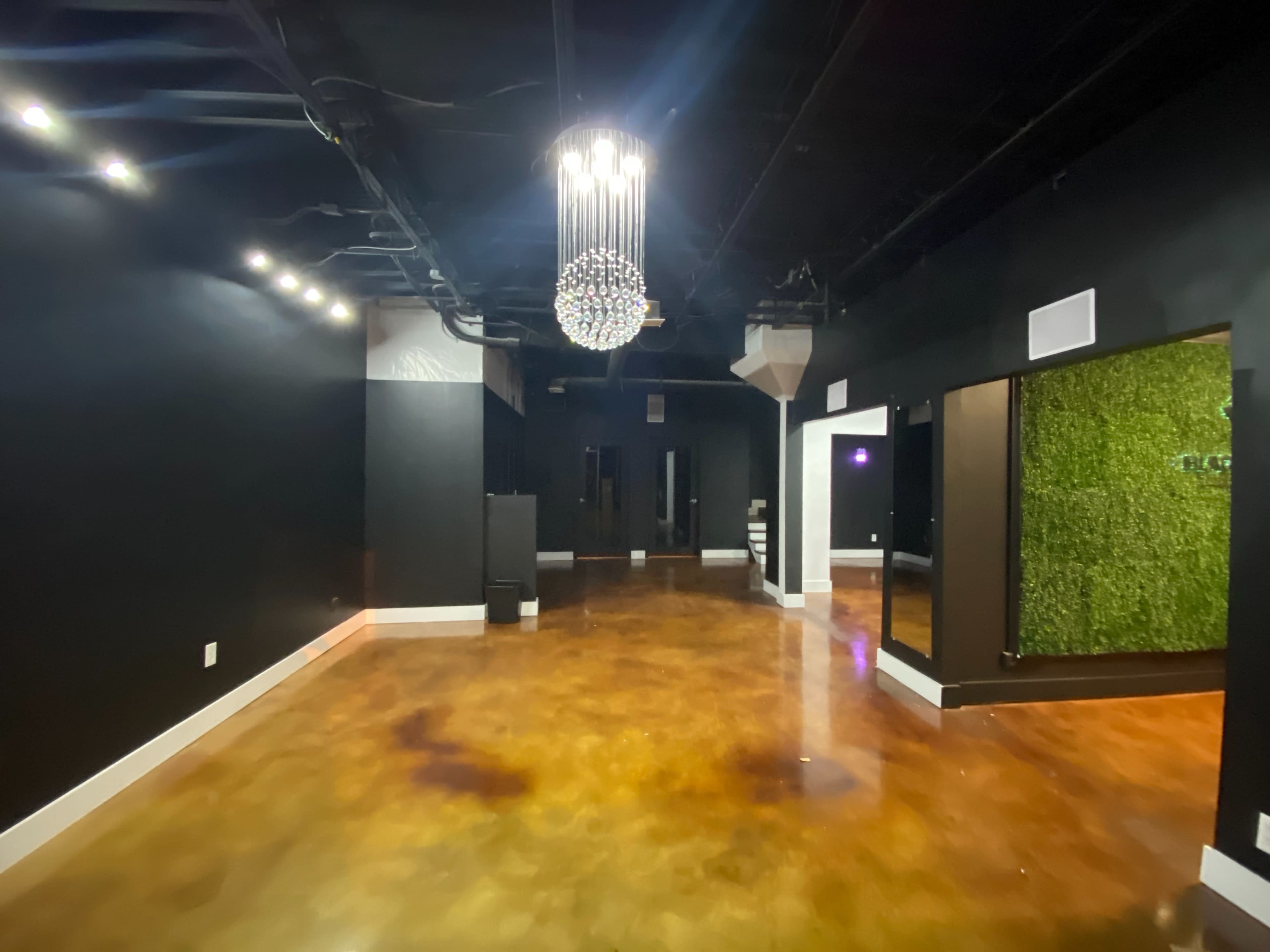 The image shows a spacious interior with polished brown floors, black walls, and a modern chandelier overhead, featuring a green wall and mirrors.