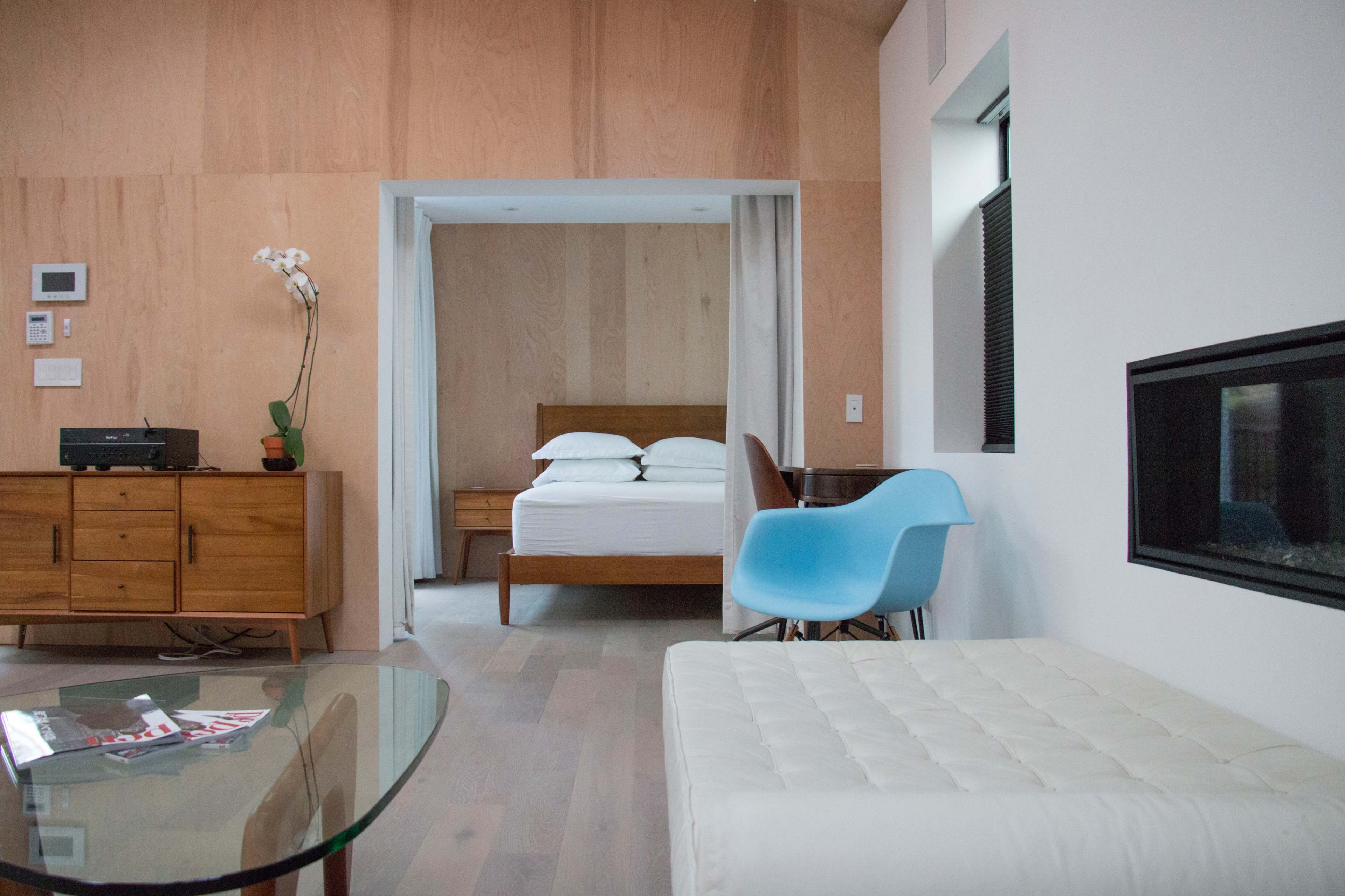The image shows a modern interior featuring a living area with a glass coffee table, a white ottoman, a blue chair, and a separate bedroom in the background with a wooden bed and nightstands.