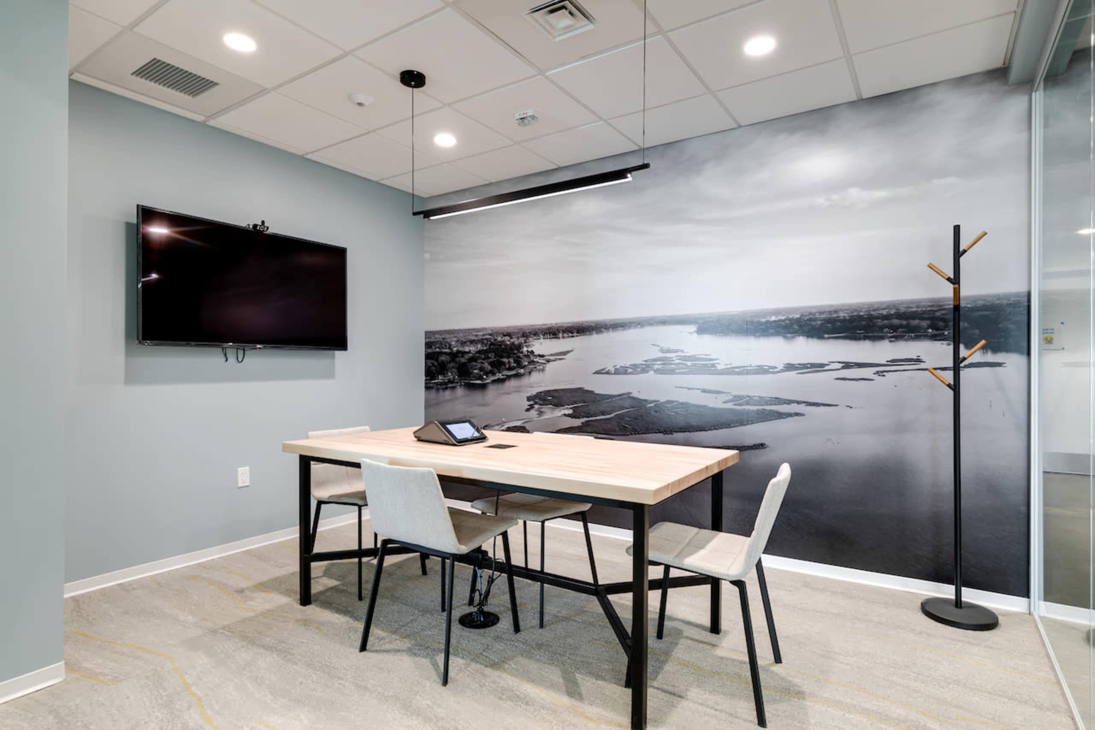 A modern conference room featuring a wooden table with four chairs, a mounted television, and a large black-and-white wall mural of a landscape.