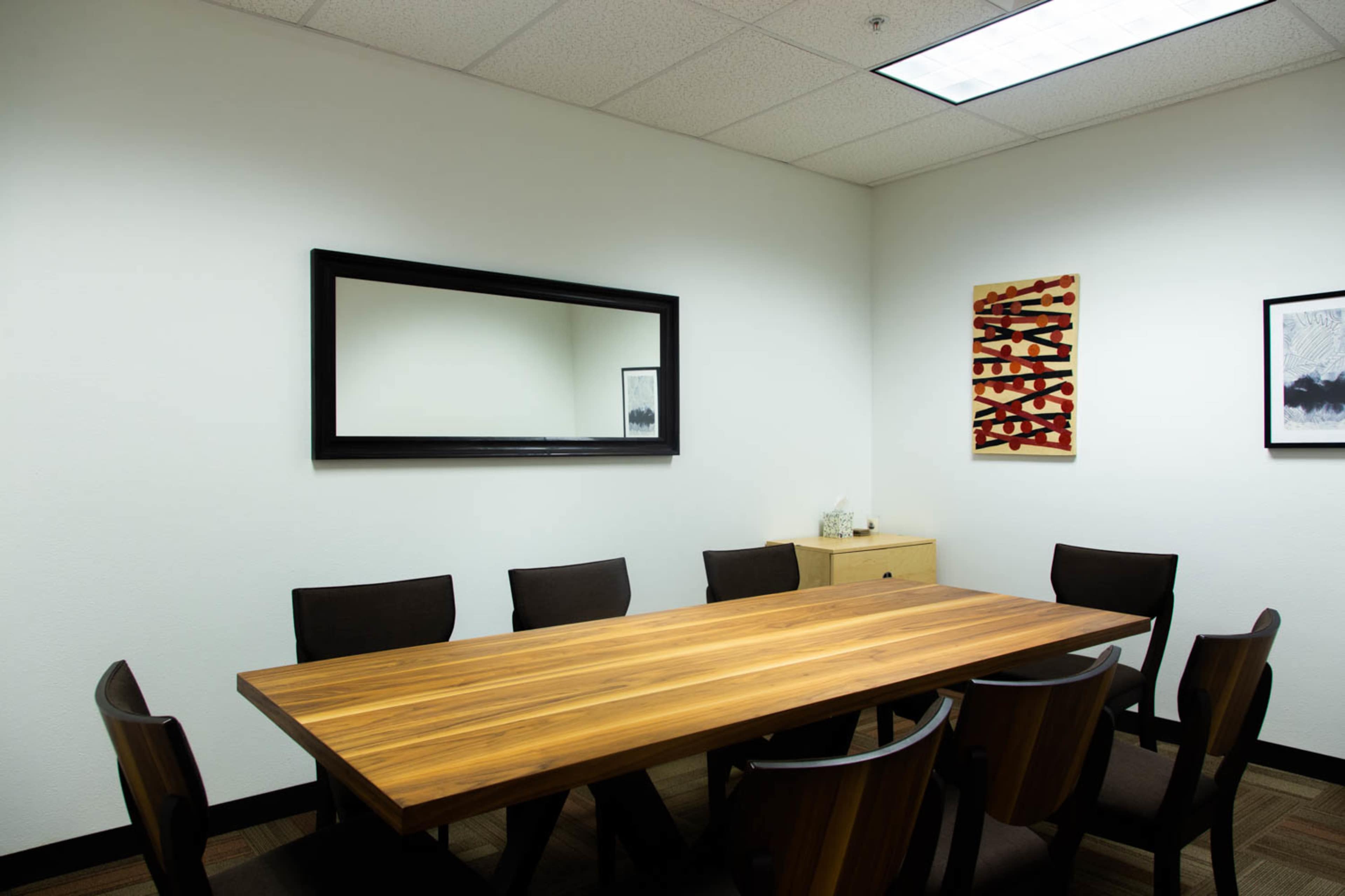 A wooden conference table with eight black chairs is set in a well-lit meeting room featuring a mirror and two decorative framed pieces on the wall.