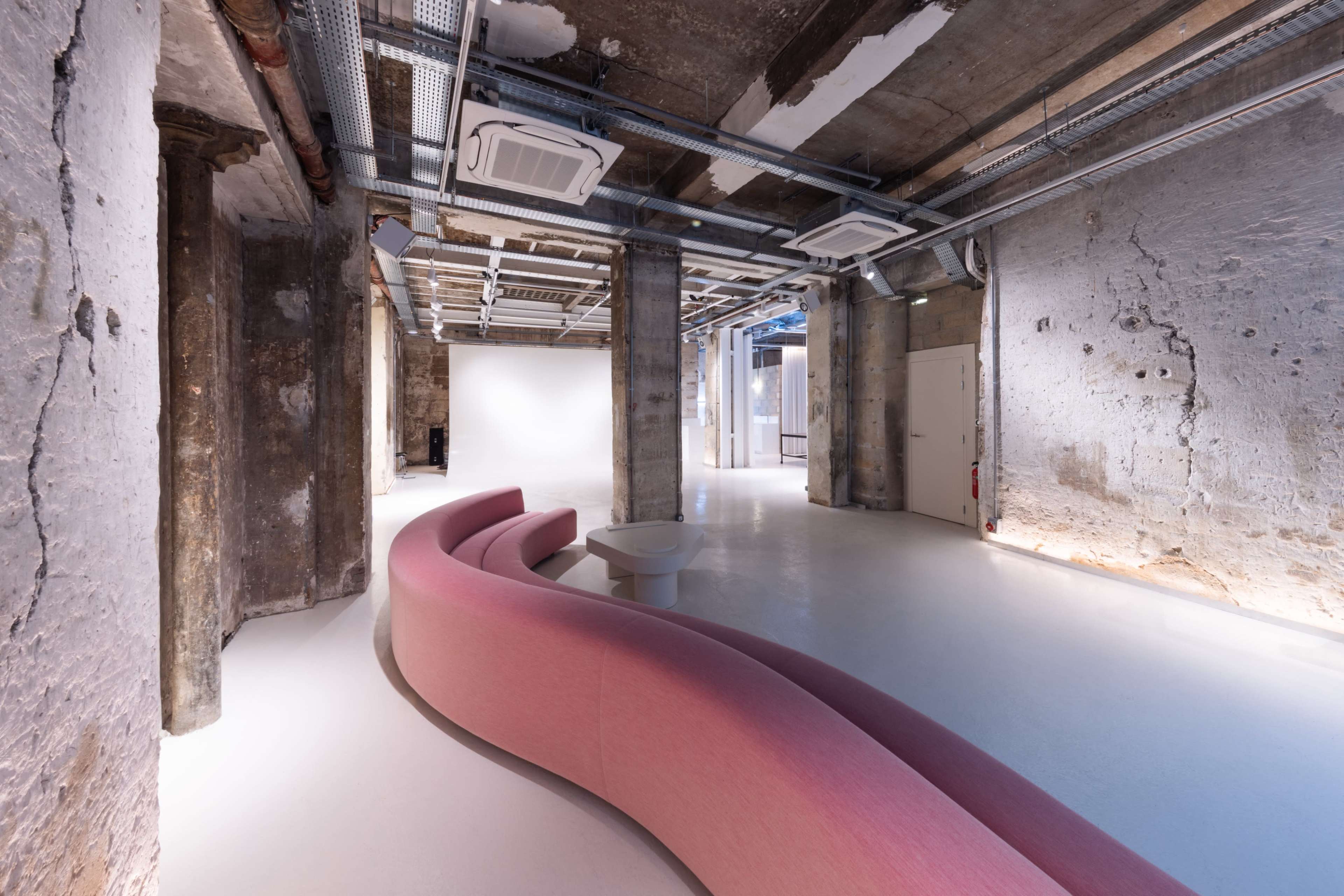 The image shows a spacious, modern interior with a curved pink sofa, exposed concrete walls, and industrial-style ceiling elements.