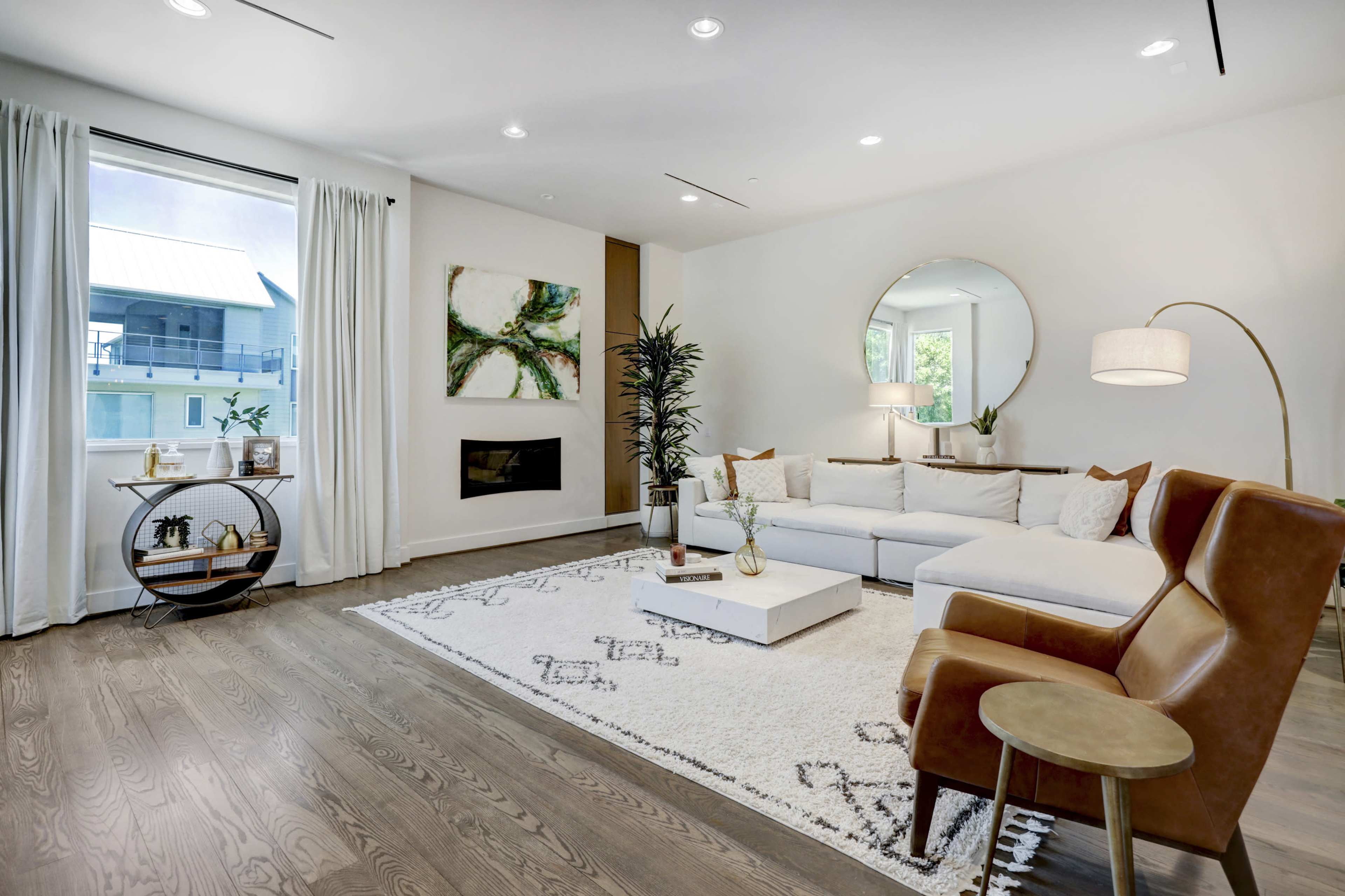 A modern living room features a white sectional sofa, a round mirror, and a wooden coffee table on a patterned rug, complemented by plants and decorative items.