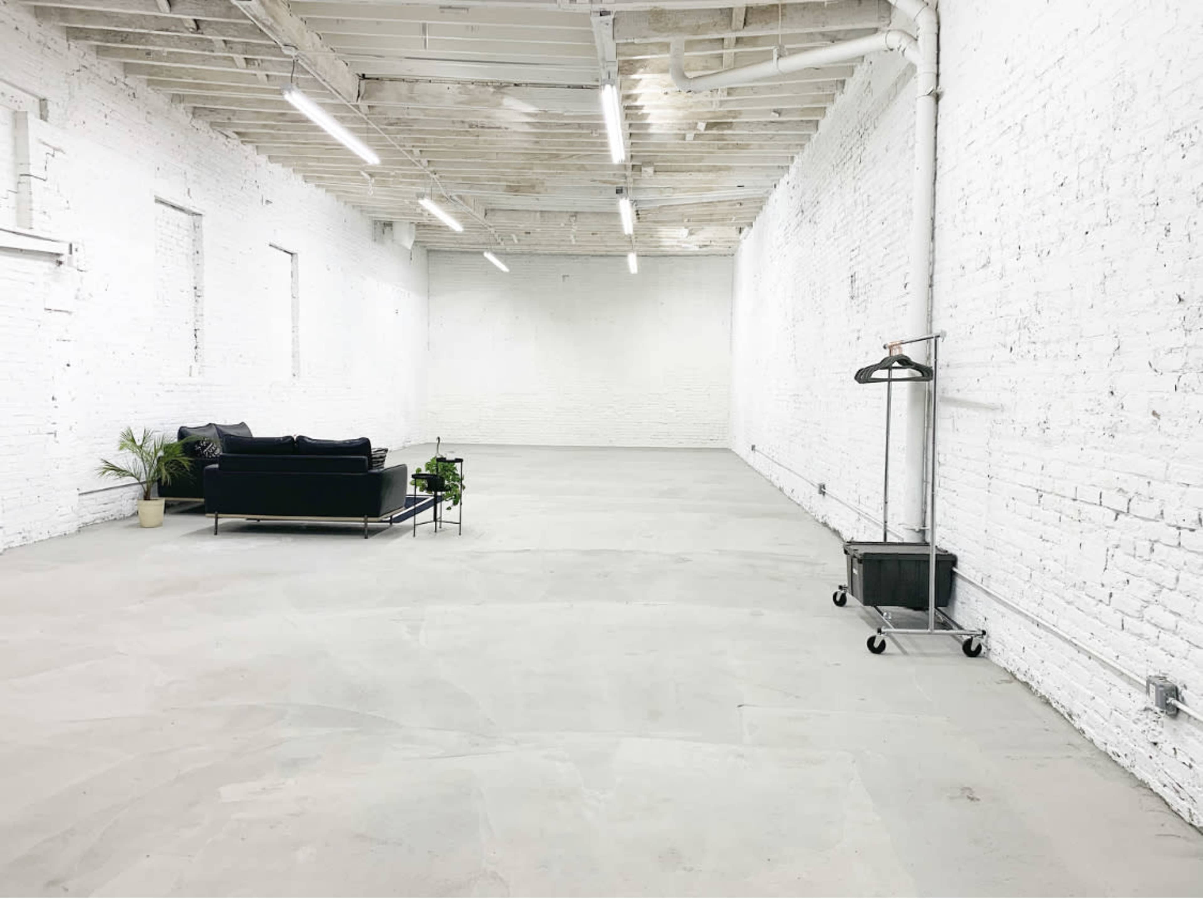 A spacious, empty room with white brick walls, a concrete floor, a black sofa, a plant, and a clothing rack on wheels.