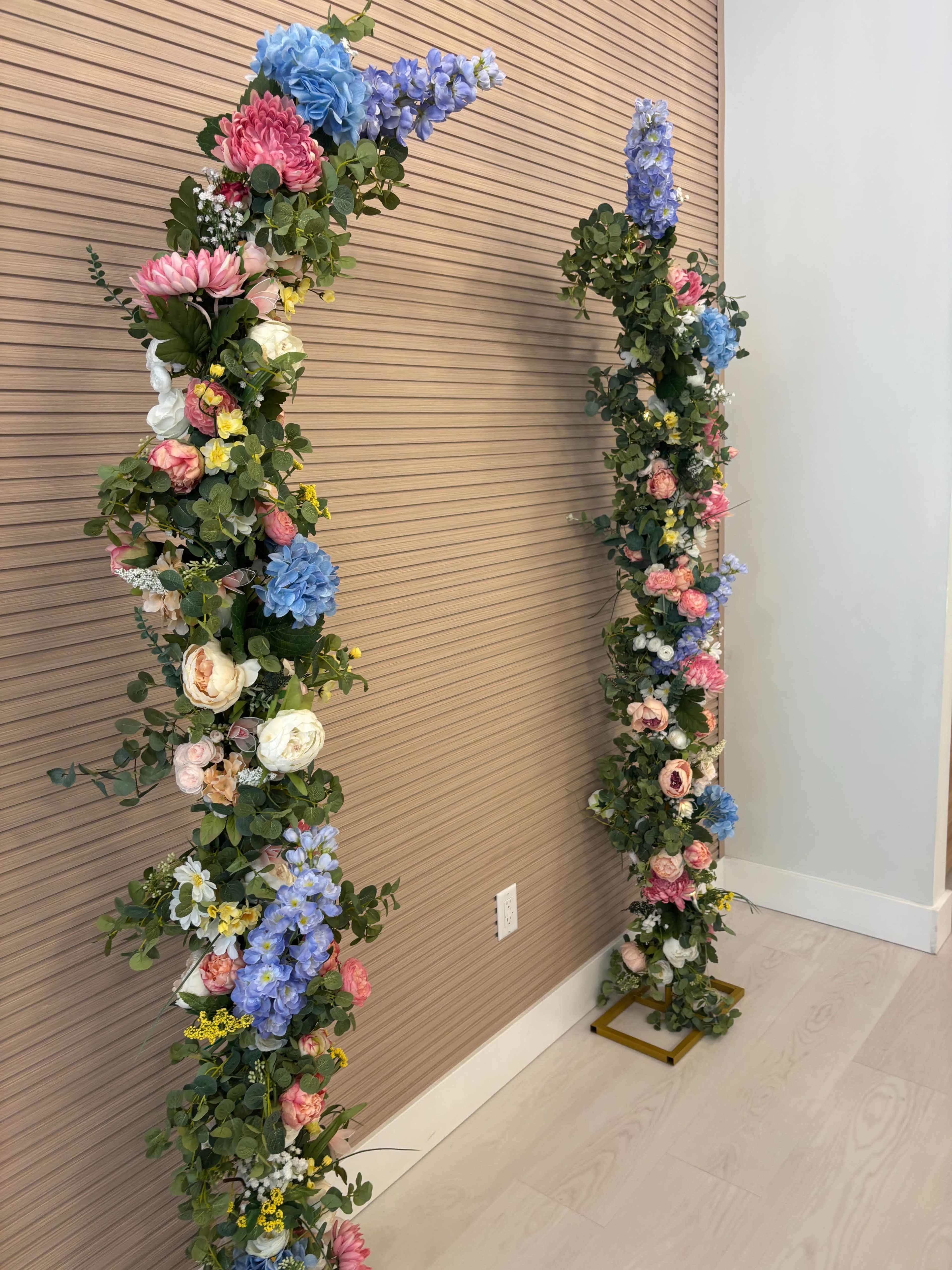 The image shows two floral arch structures adorned with a variety of artificial flowers against a wall with horizontal wood paneling.