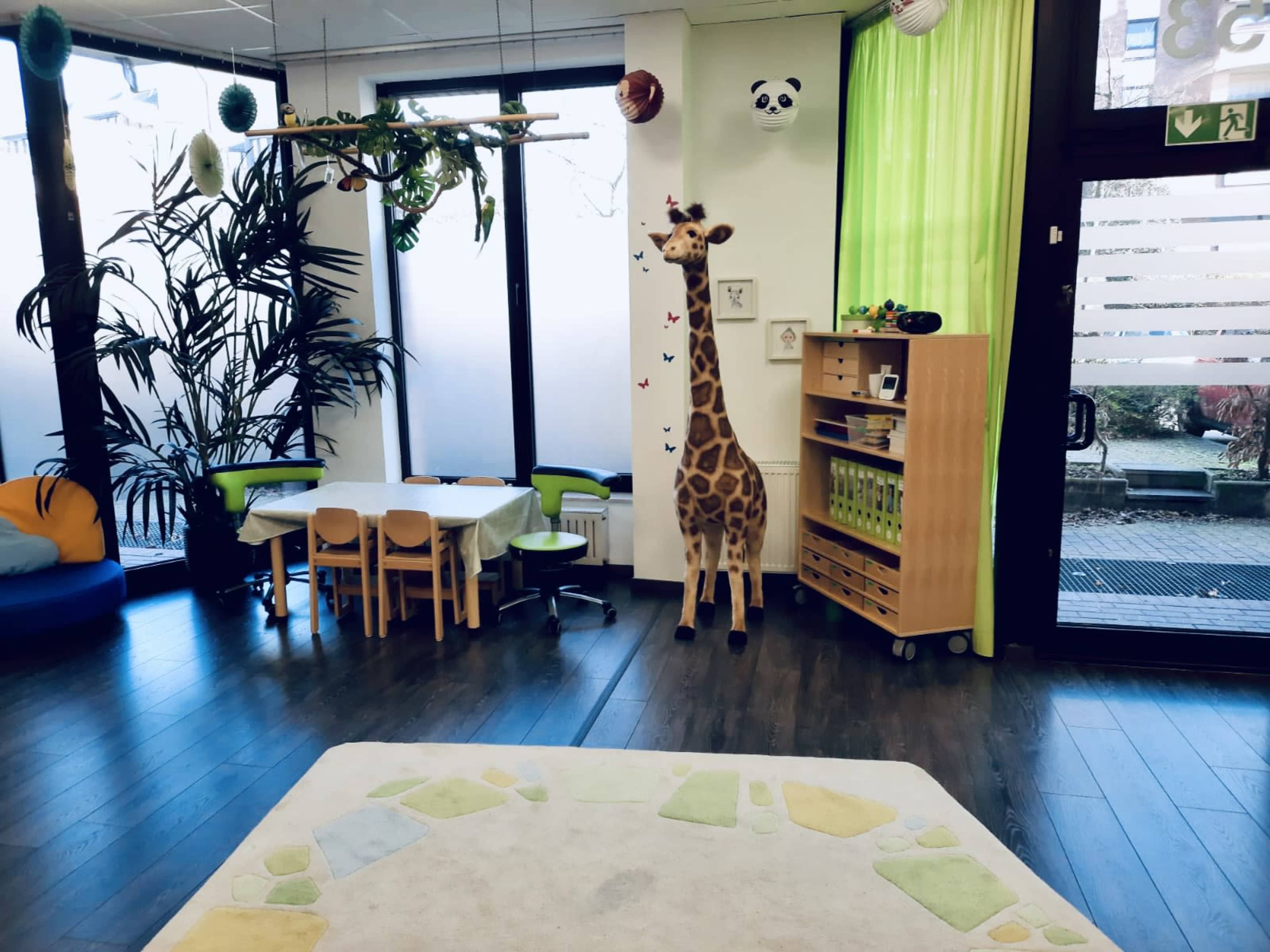 The image shows a child-friendly room featuring a small table and chairs, a bookshelf, plants, and a giraffe decoration against a bright green wall.