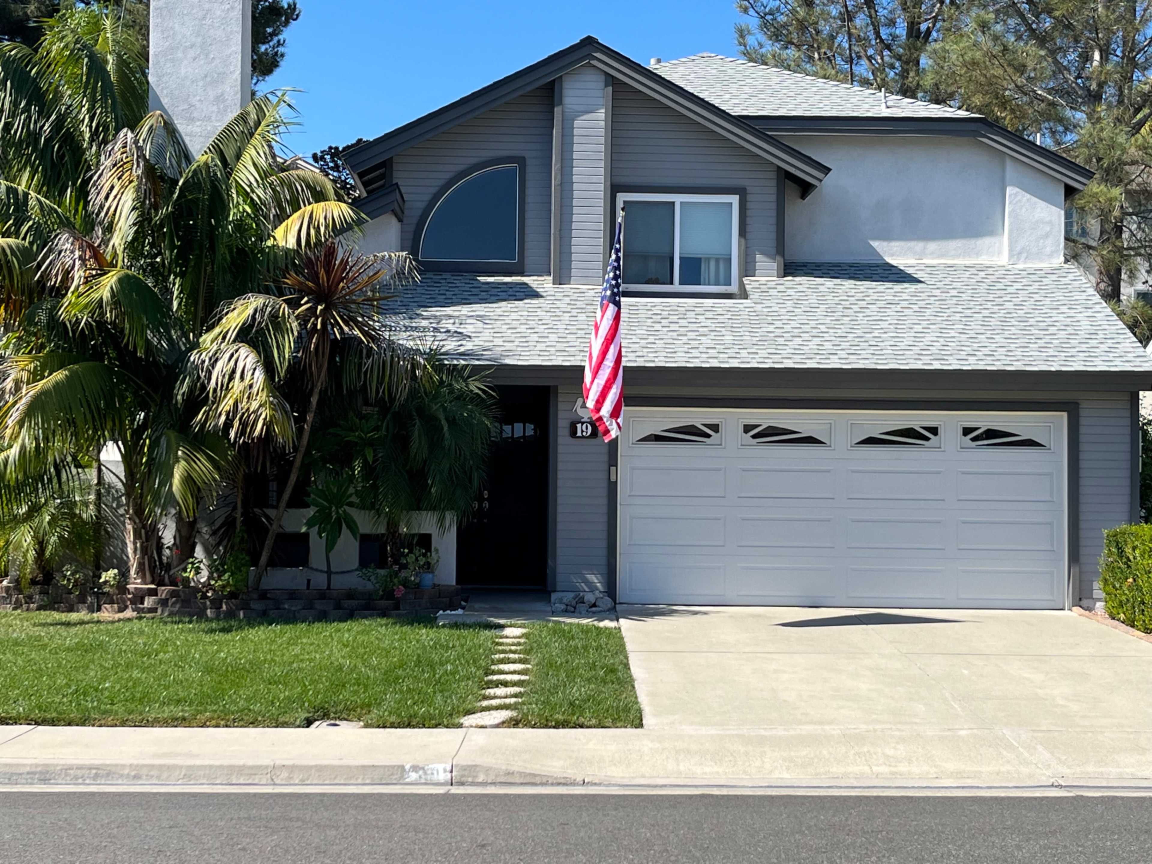 A two-story gray house with a double garage, an American flag hanging from the porch, and landscaped greenery in the front yard.