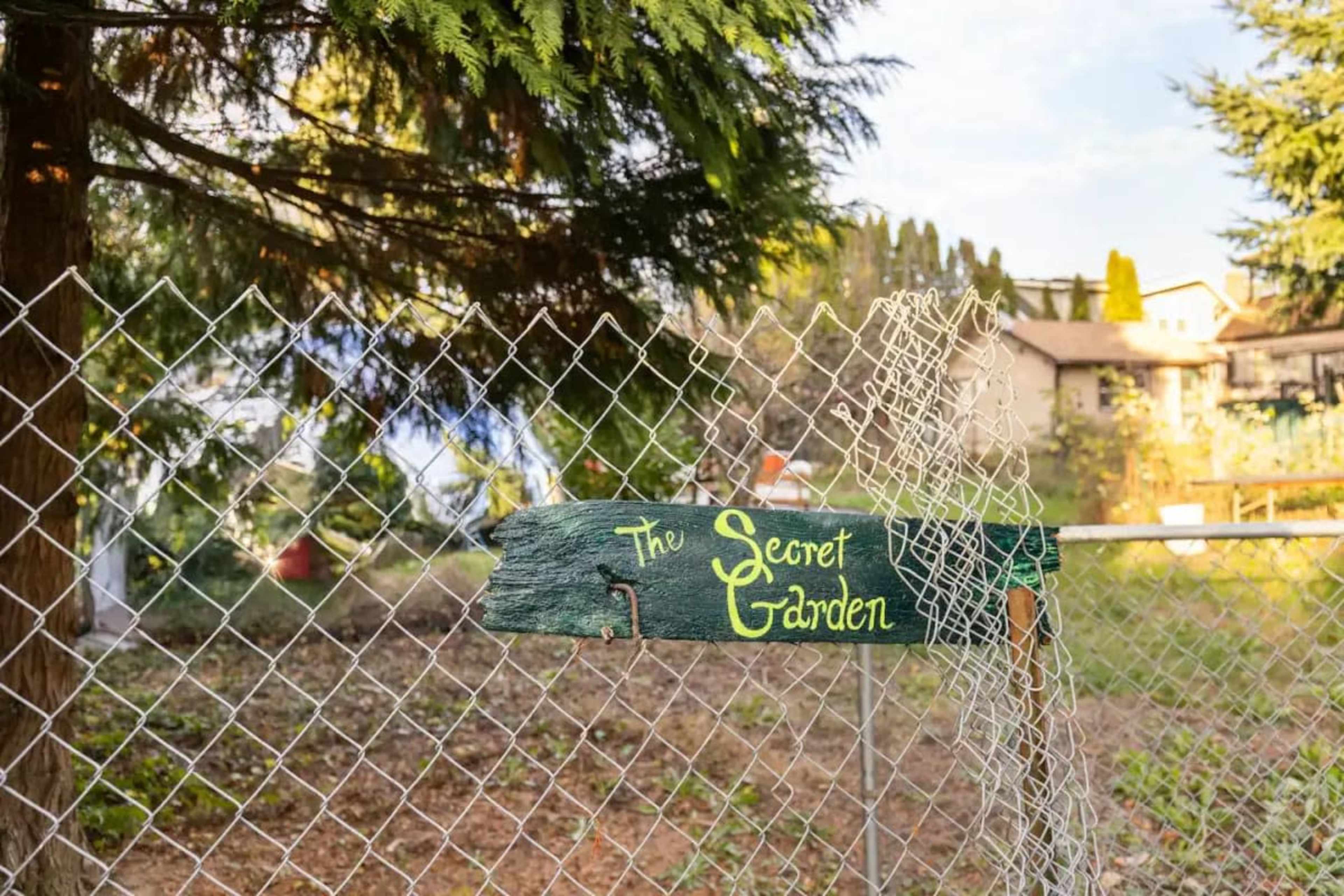 A wooden sign reading "The Secret Garden" hangs on a chain-link fence surrounded by trees and a garden area.