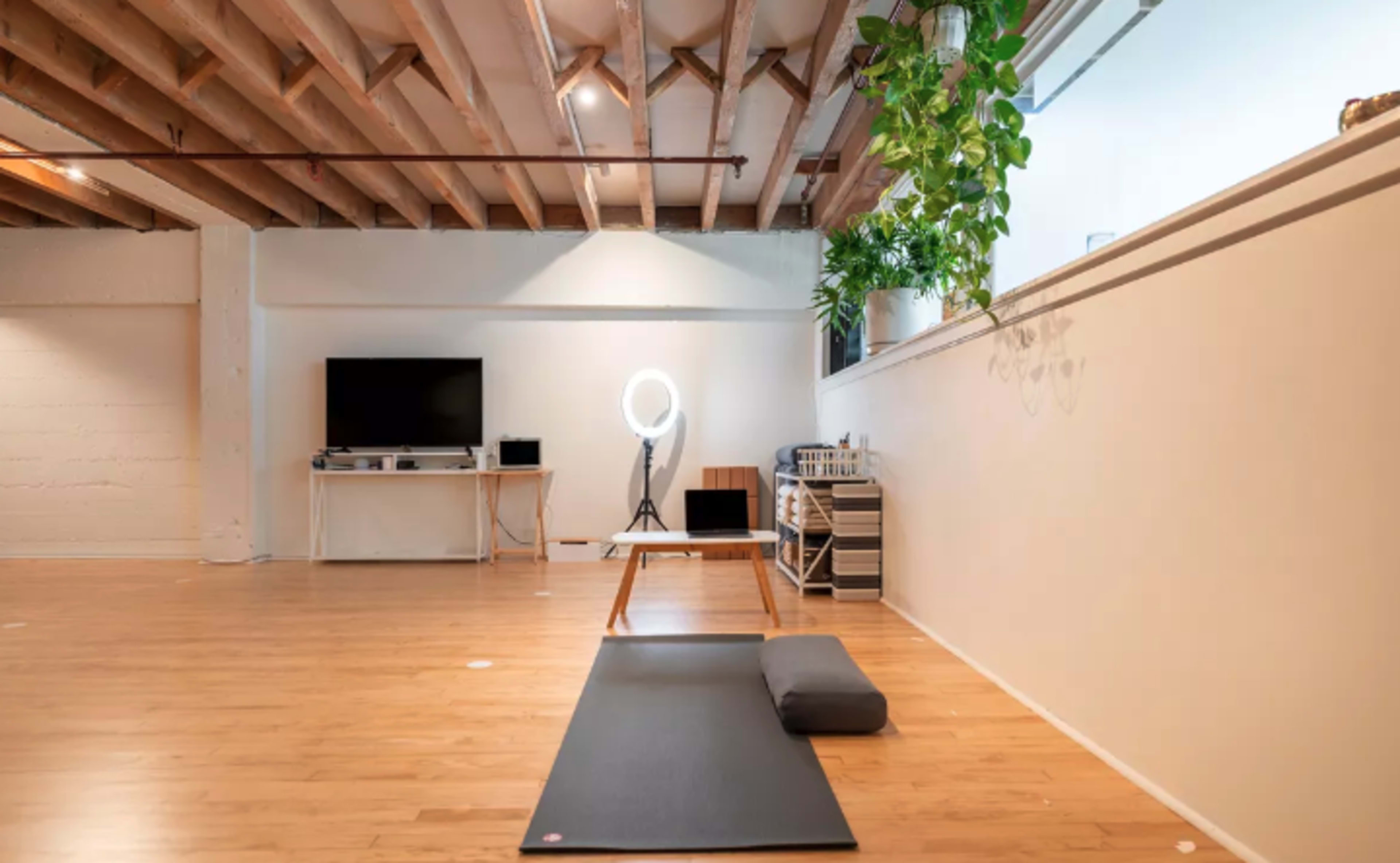 A spacious, minimalistic yoga studio with wooden flooring, a yoga mat, a cushion, and equipment set against a white wall.