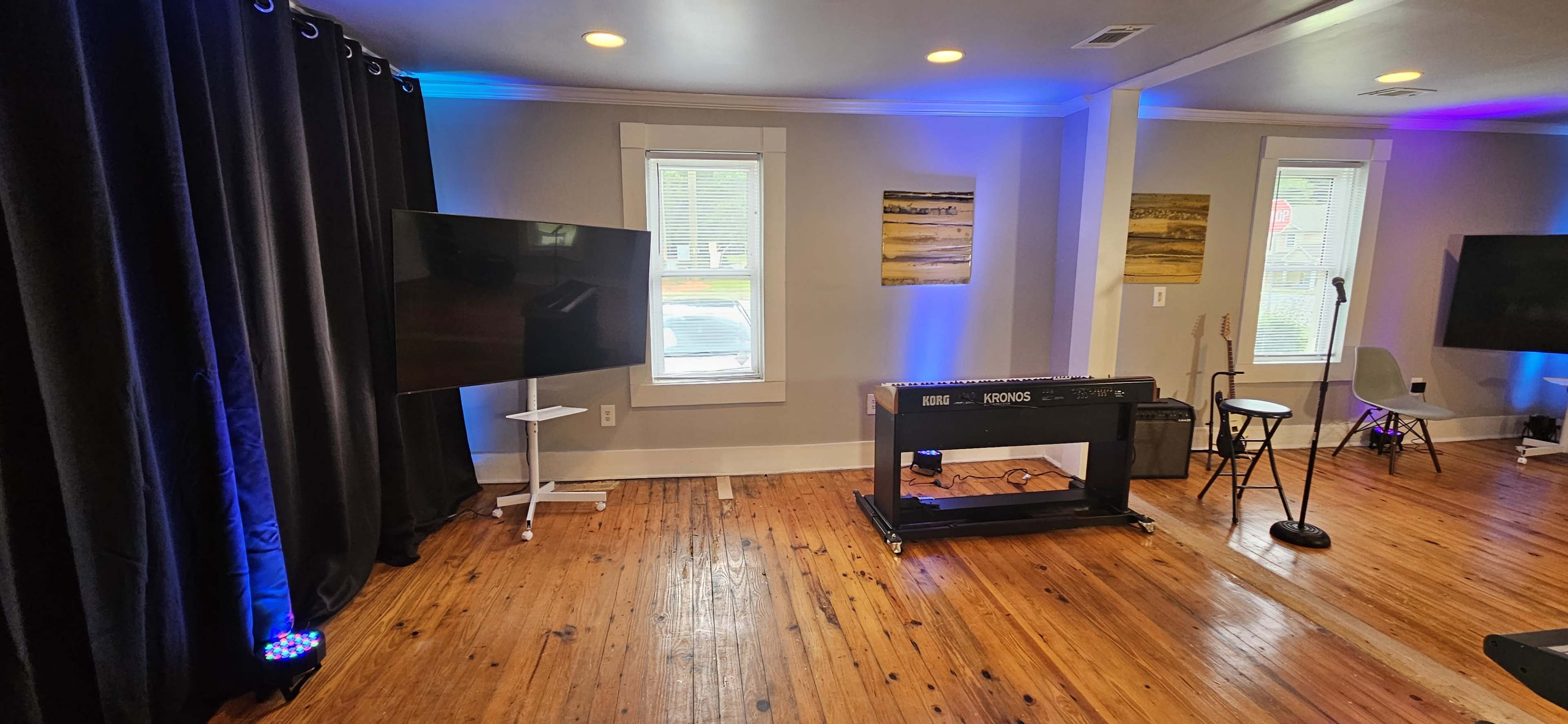 The room features a keyboard on a stand, two televisions, and a microphone stand, all arranged with blue lighting and wooden floors.