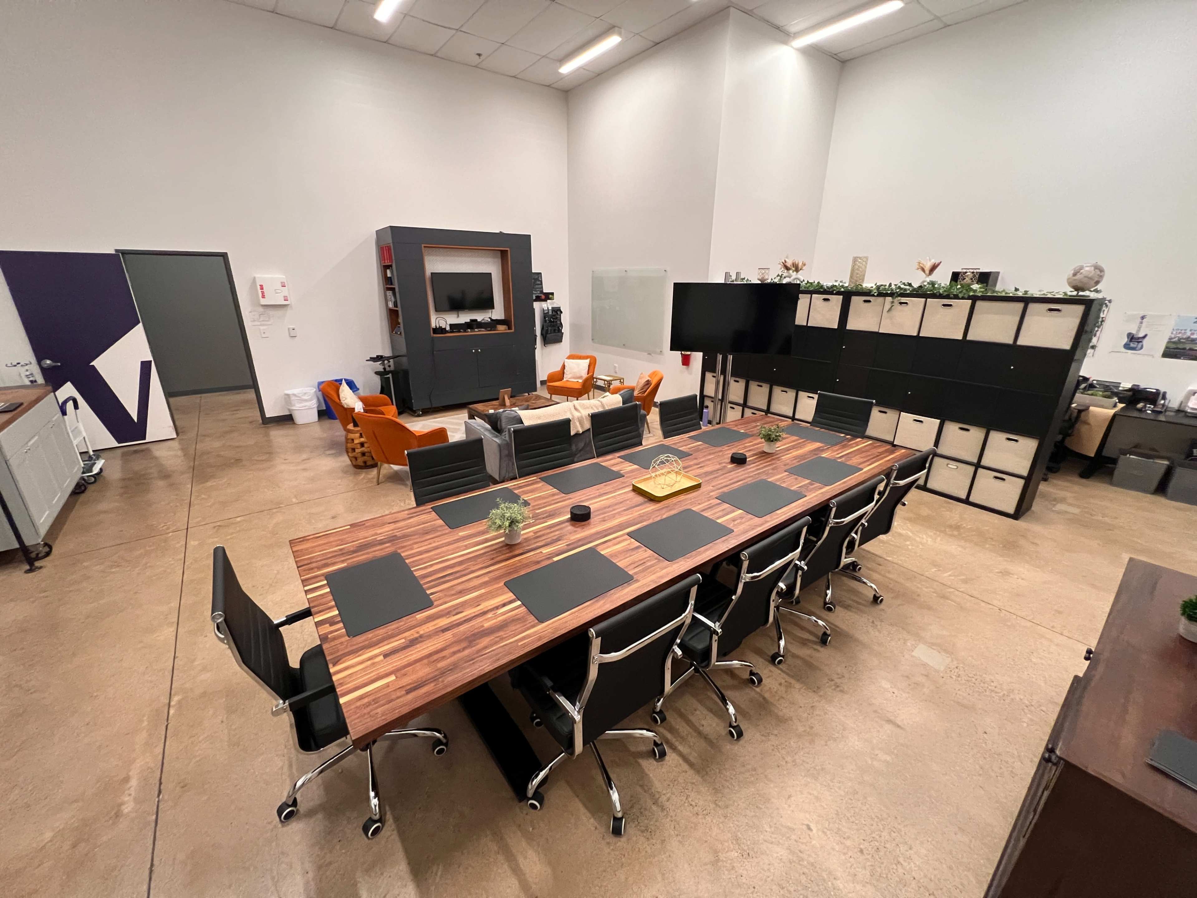 The image shows a modern conference room featuring a large wooden table surrounded by black office chairs and separate seating areas with orange chairs.