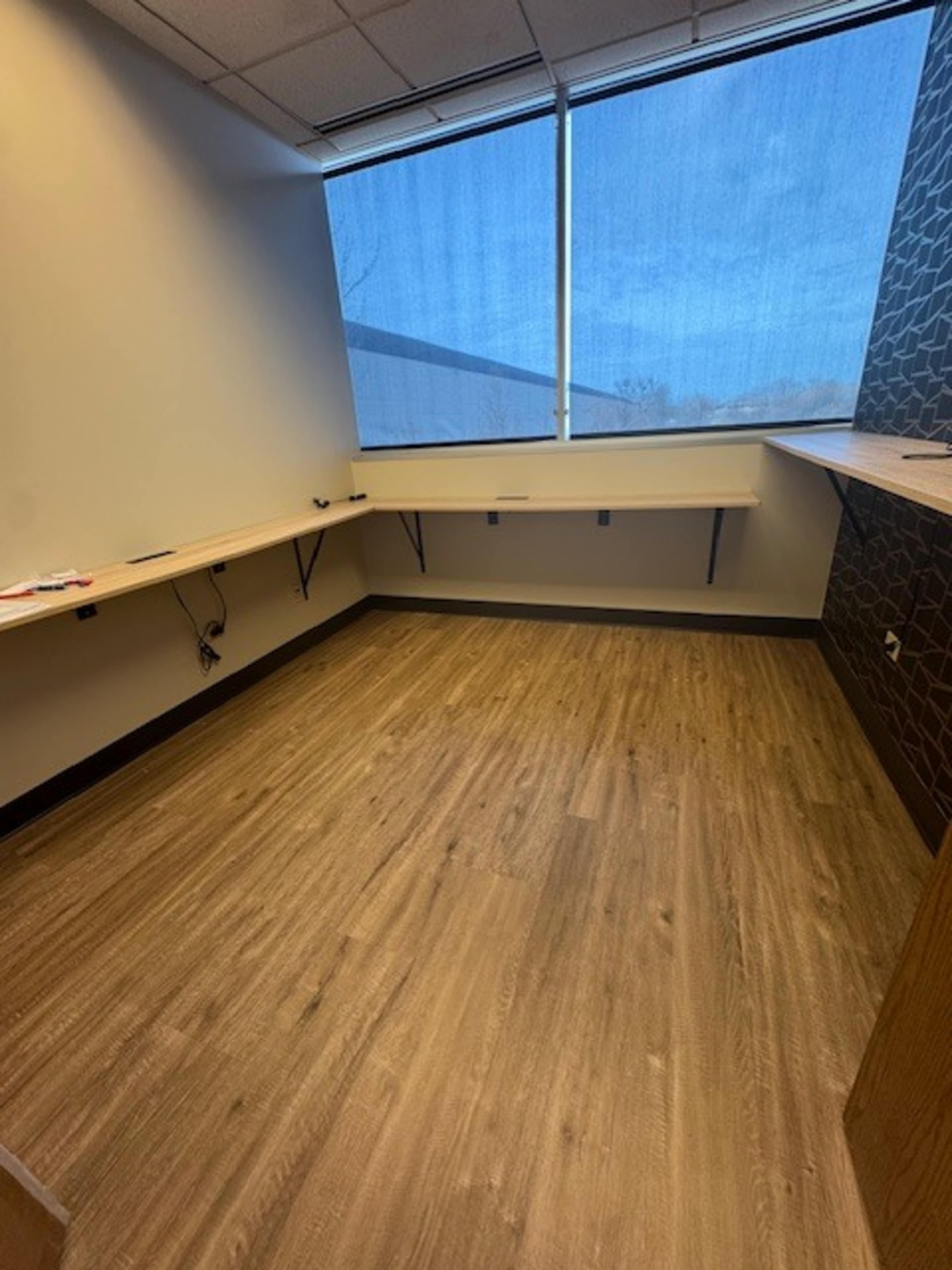 The image shows an empty office space with wooden flooring, two long shelves attached to the walls, and a large window.