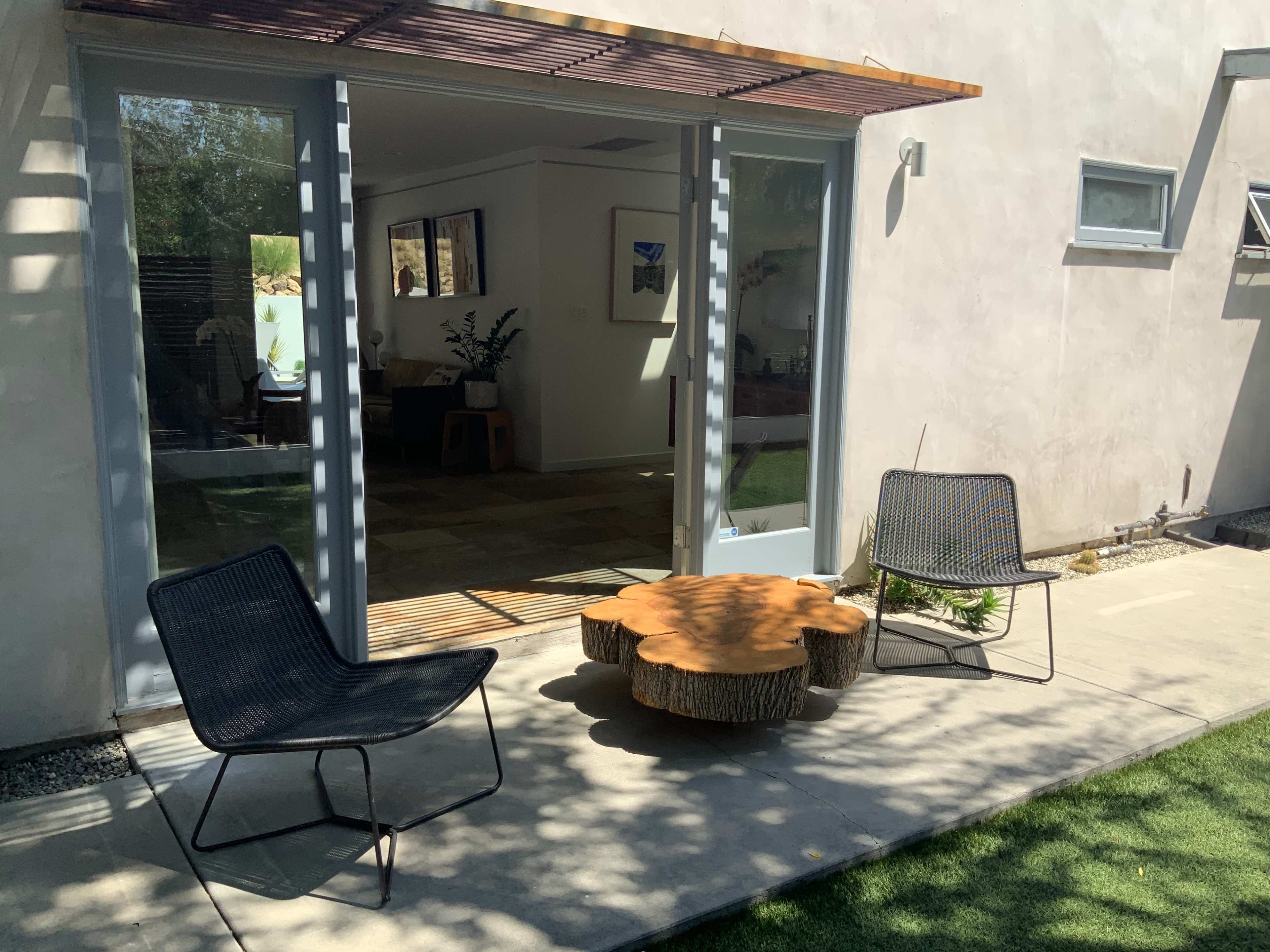 The image shows a small outdoor seating area with two black chairs and a round wooden table in front of a house with large glass doors.
