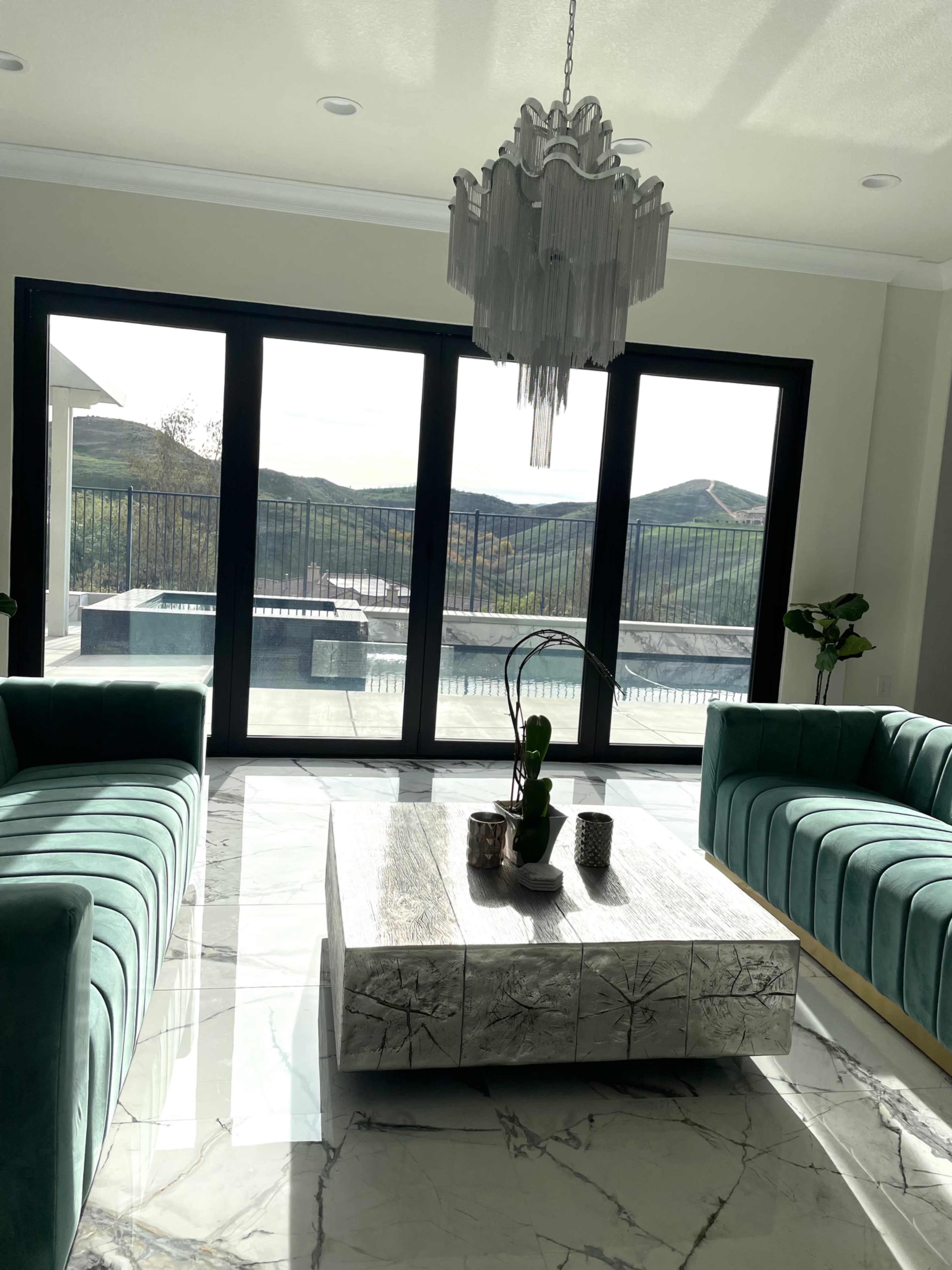 A modern living room with two teal sofas facing a glass coffee table, large windows providing views of a pool and surrounding hills.