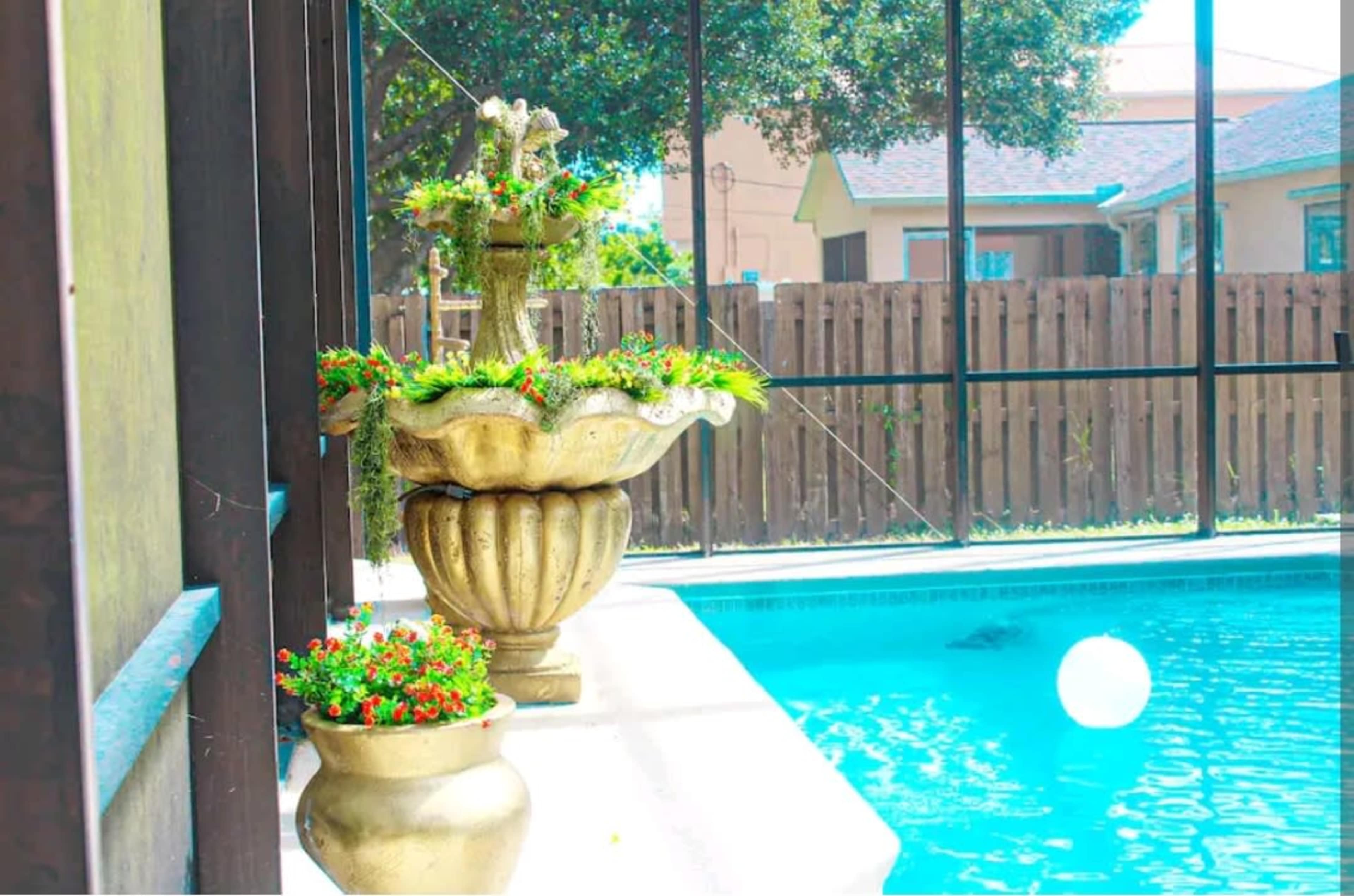 A decorative fountain with potted plants beside a swimming pool enclosed by a screened-in patio.