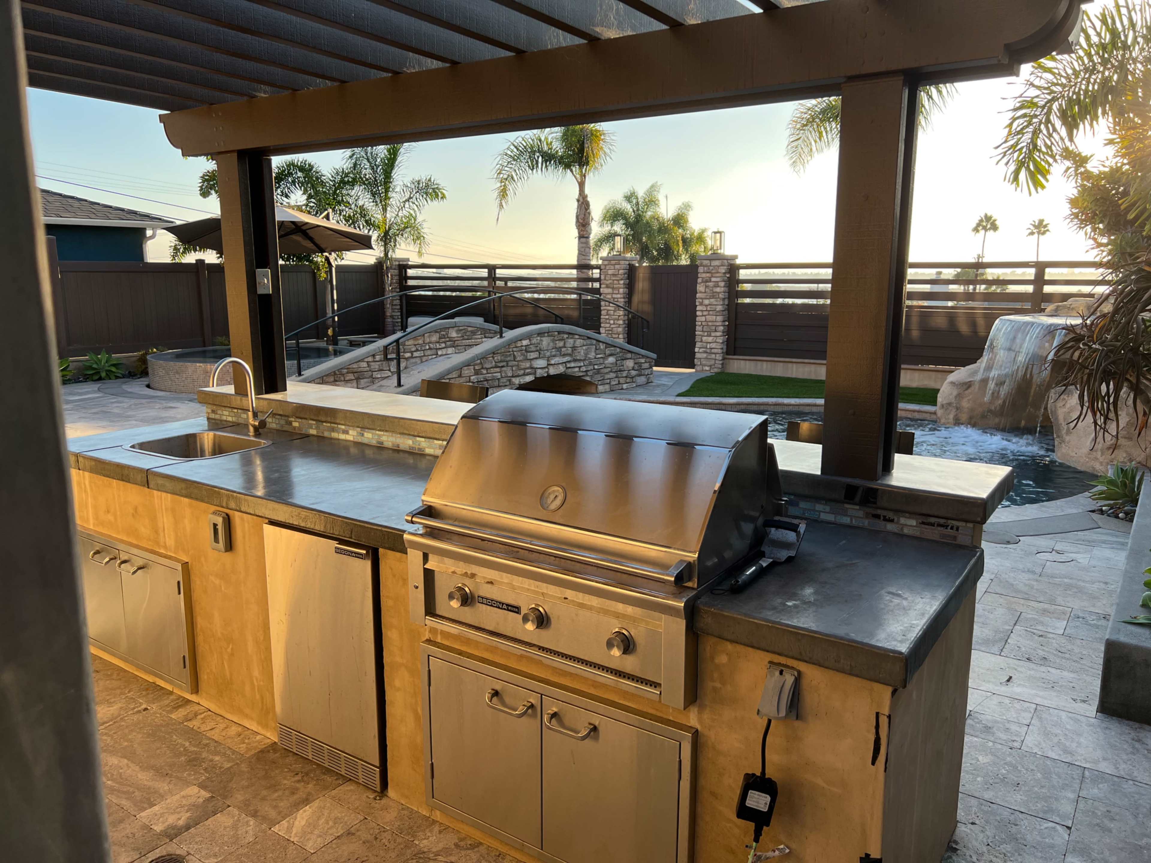 An outdoor kitchen with a stainless steel grill, sink, and countertop, surrounded by palm trees and a landscaped backyard.