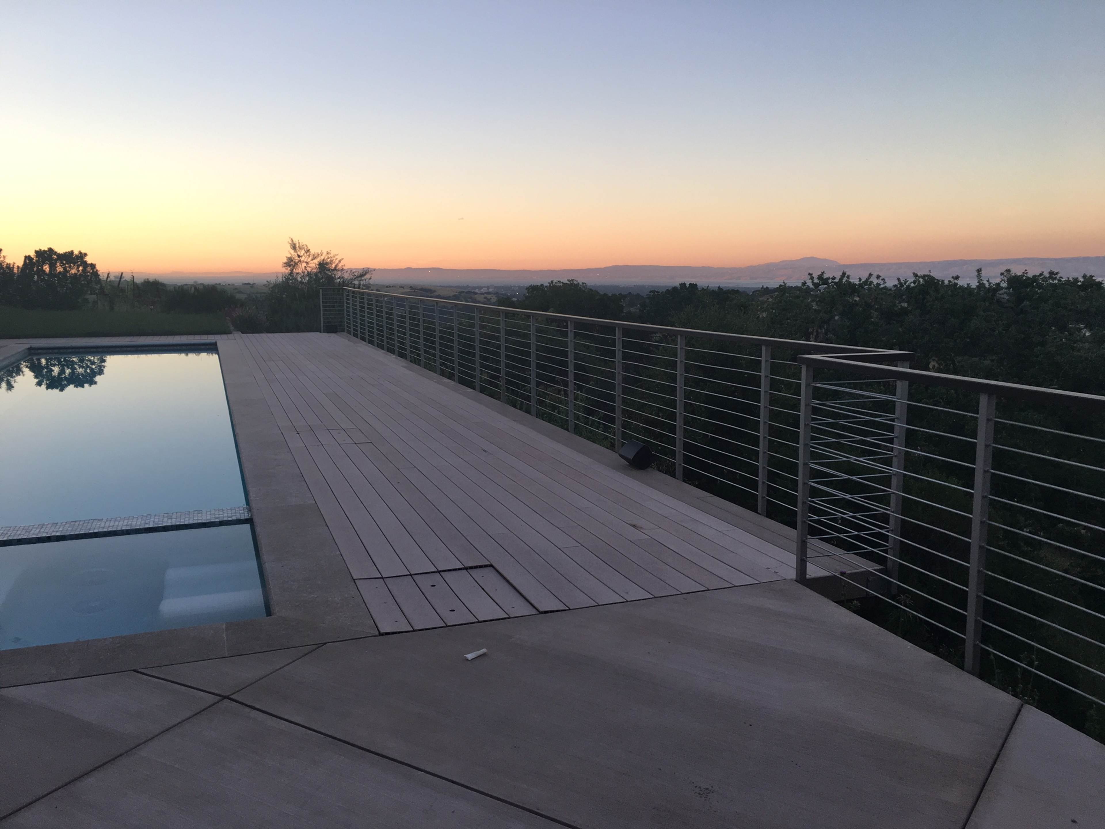 A wooden deck with a railing overlooks a pool and a distant landscape at sunset.