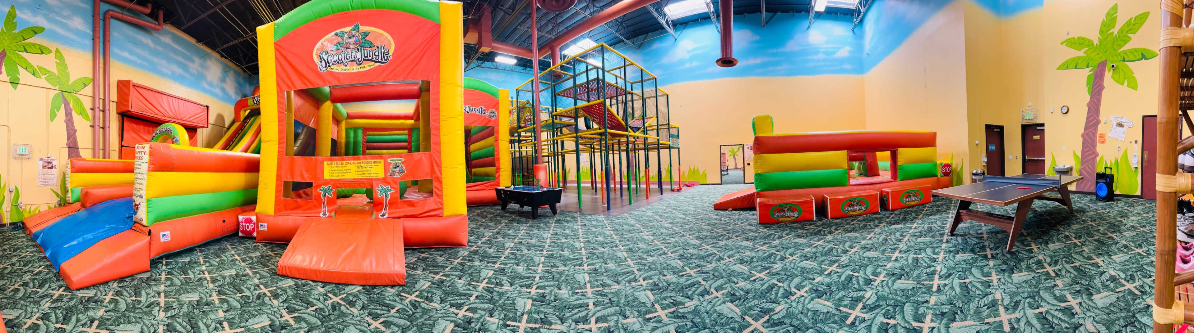 The image shows a brightly colored indoor play area featuring inflatable structures, a climbing frame, and a ping pong table against a painted backdrop of palm trees and a blue sky.