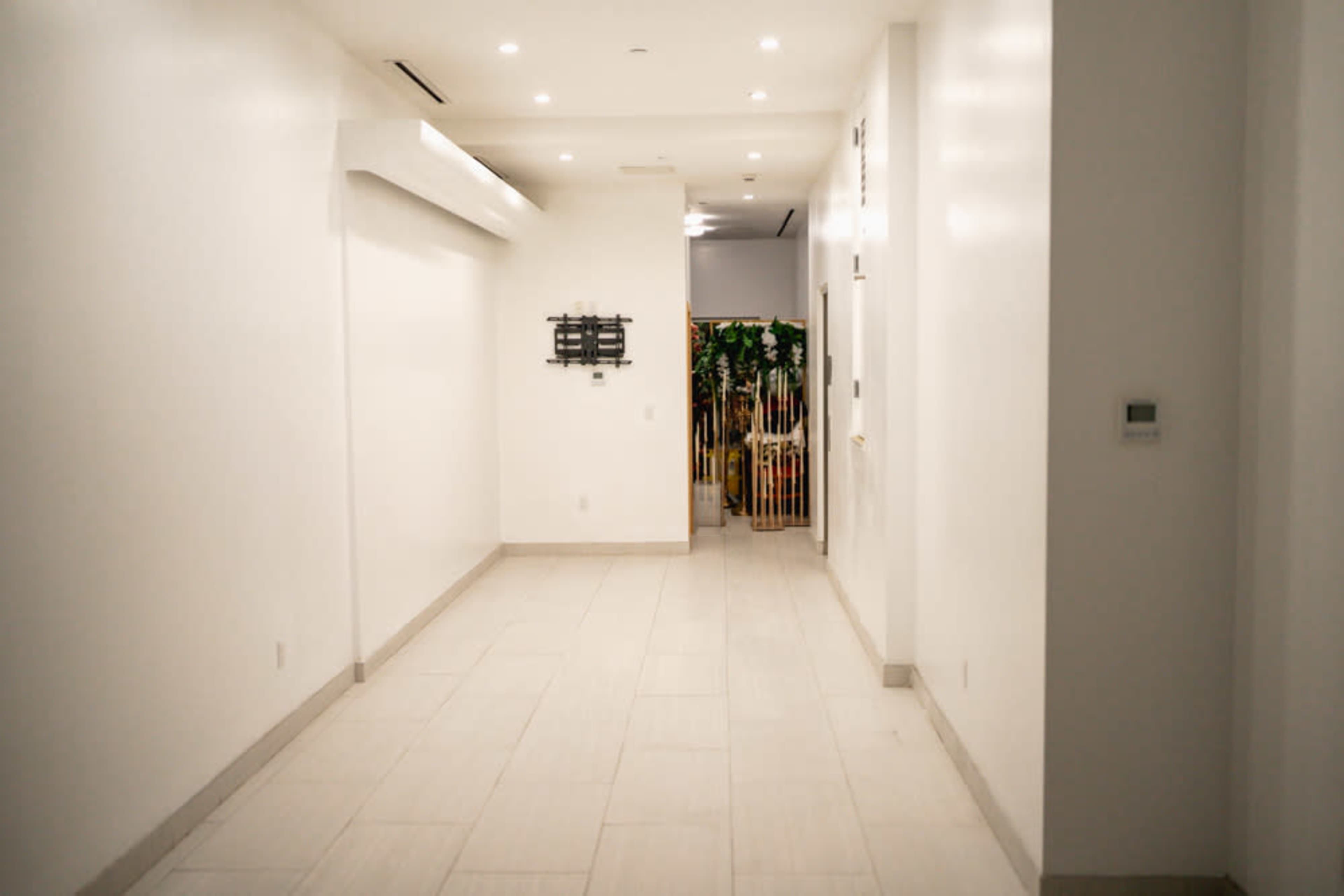 The image shows a long, white hallway with polished tile flooring and illuminated ceiling lights, leading to a partially visible area with greenery in the background.