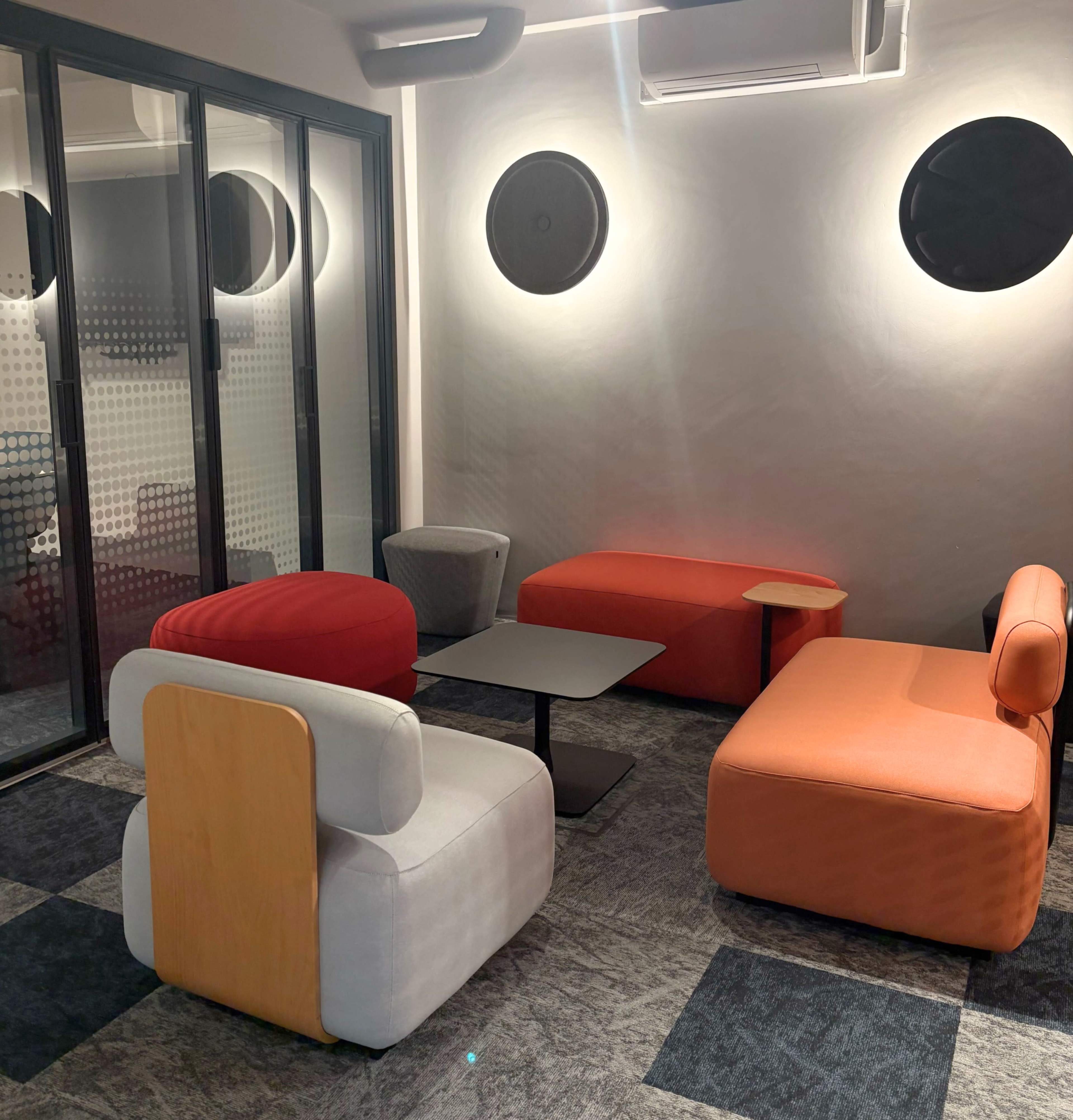 The image shows a modern lounge area with various seating options in red, orange, gray, and black, accompanied by a black table.