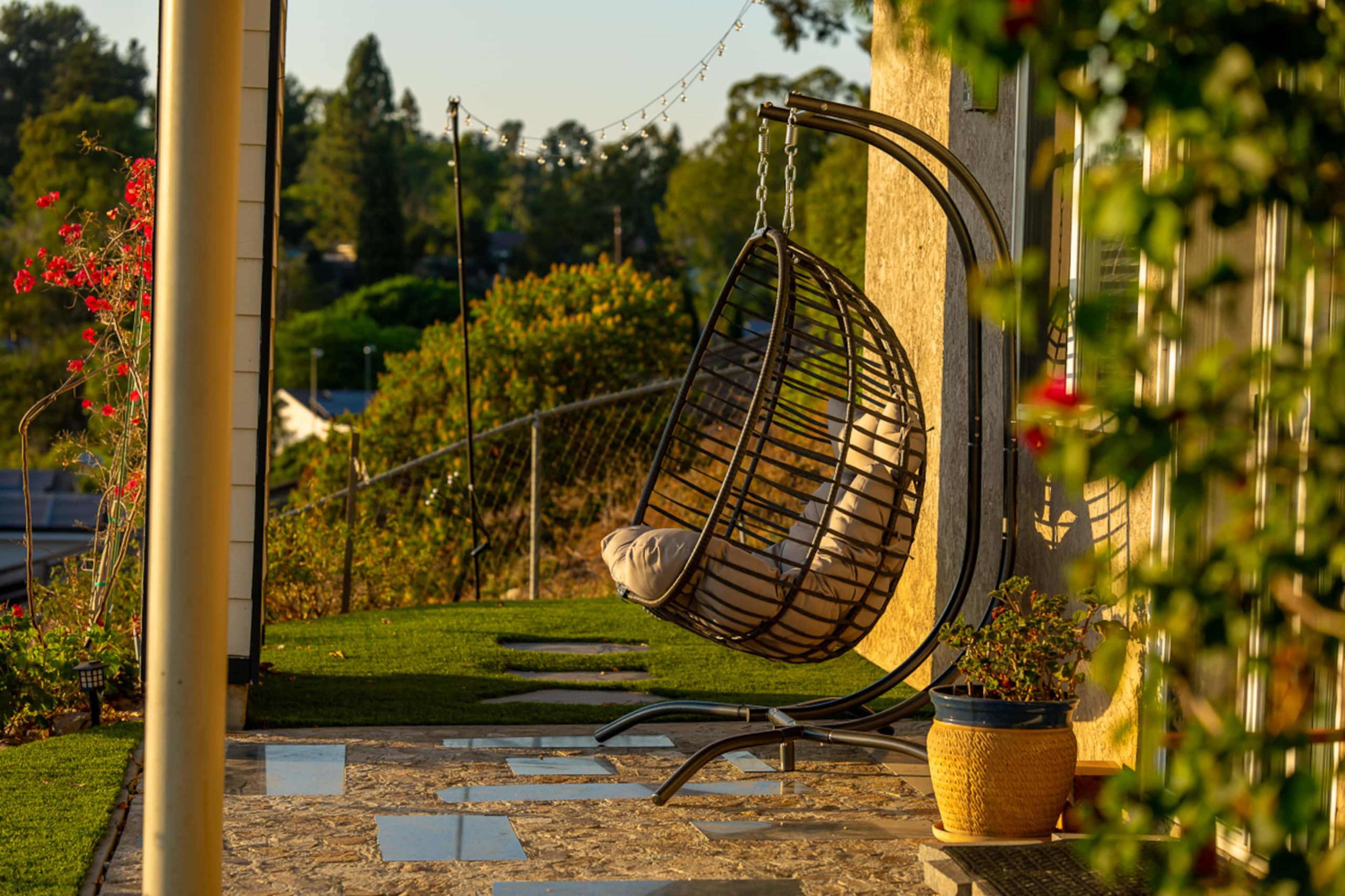 A large hanging chair is positioned on a stone patio beside a house, surrounded by greenery and plants.