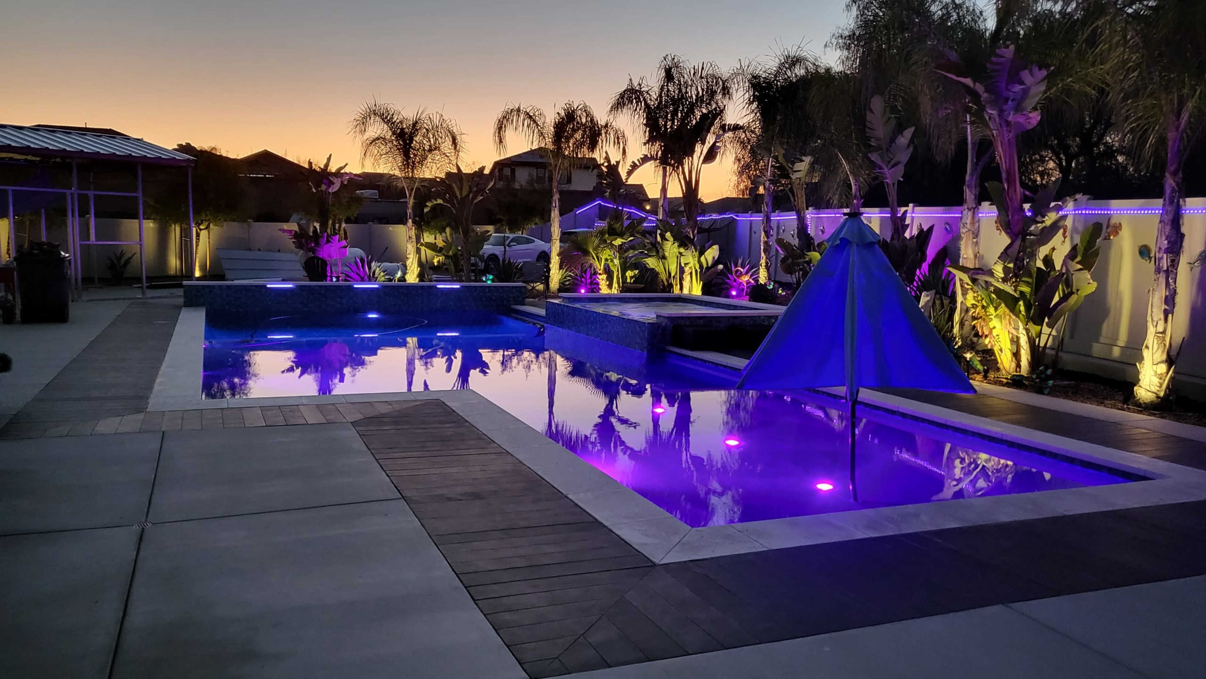 A backyard pool area features illuminated water and plants, with a triangle canopy set up beside the pool at dusk.