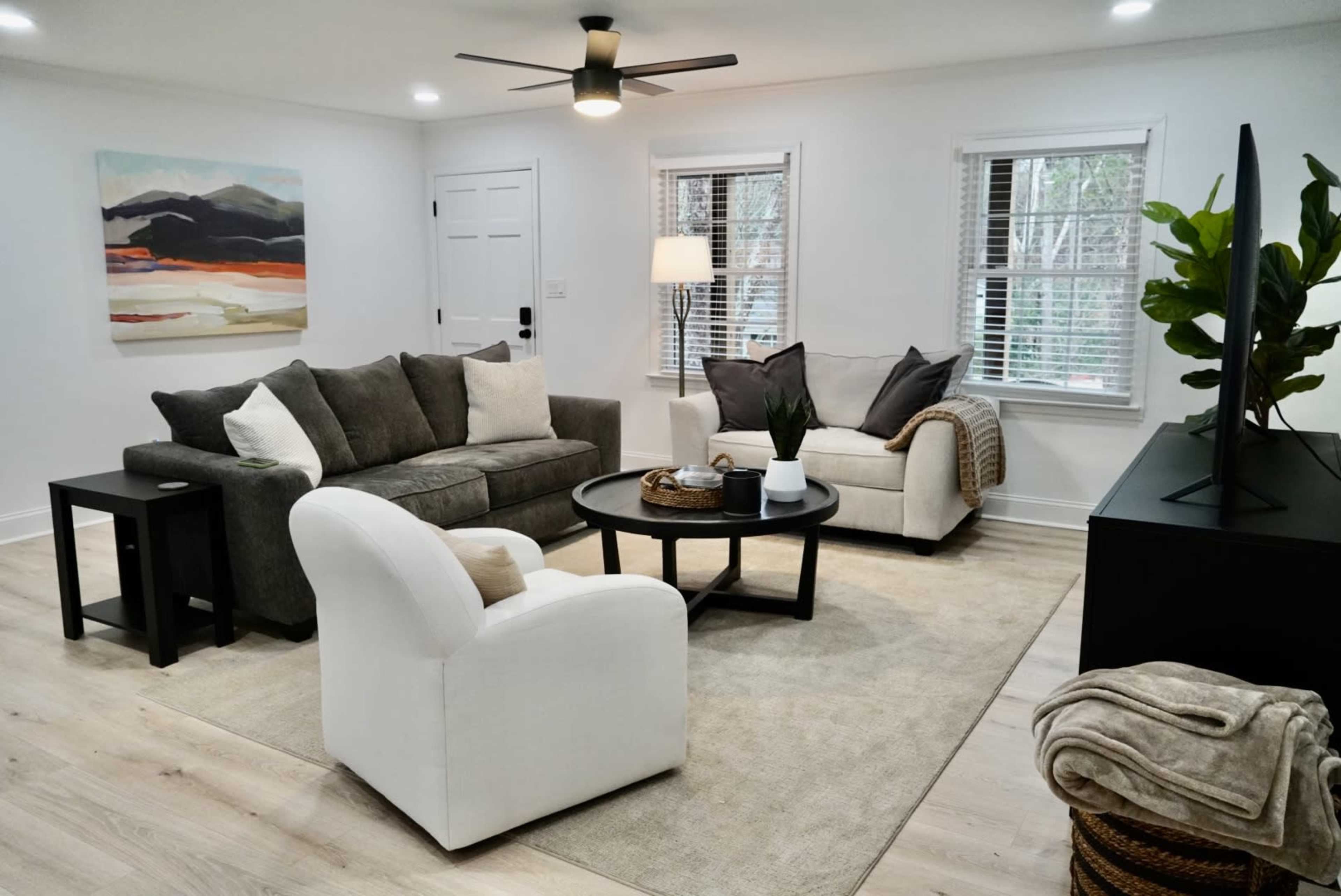 A modern living room featuring a gray sofa, a white armchair, a black coffee table, and a large television against a wall, with large windows providing natural light.
