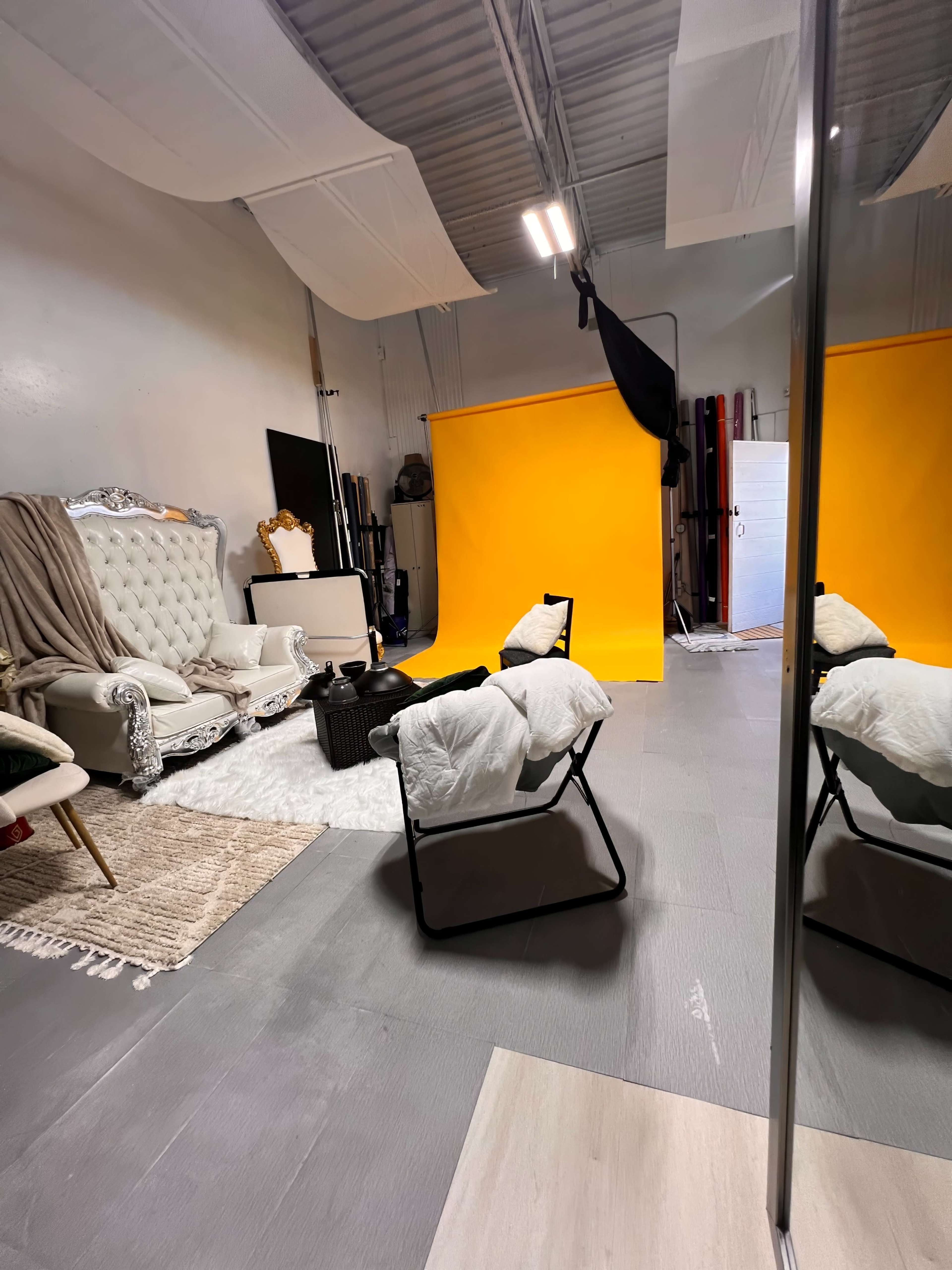 A photo studio with a yellow backdrop, a large cushioned chair, a small table, and an area rug on the floor.