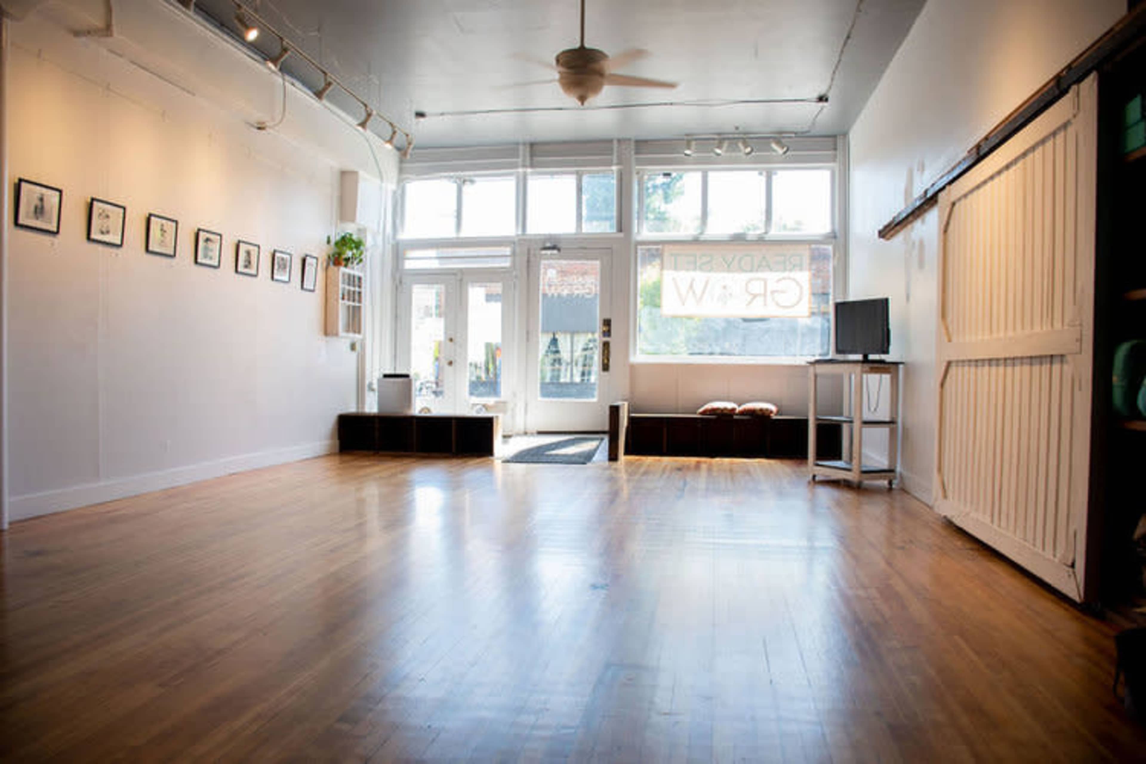 A spacious, brightly lit room with wooden flooring, featuring a few art frames on the wall and large windows letting in natural light.