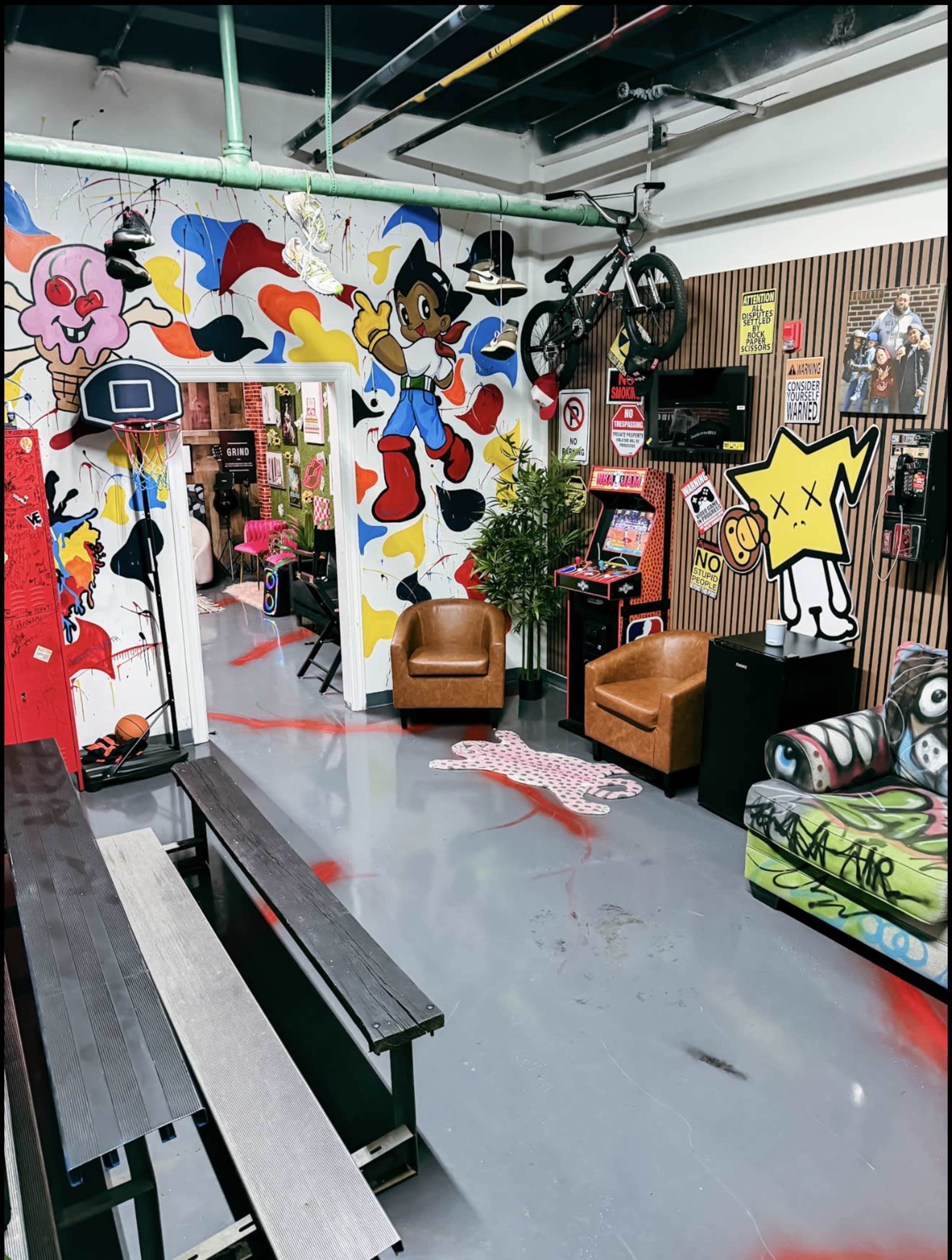 The image shows an indoor space with colorful graffiti on the walls, two brown chairs, a bicycle hanging from the ceiling, and various gaming and entertainment items scattered throughout.
