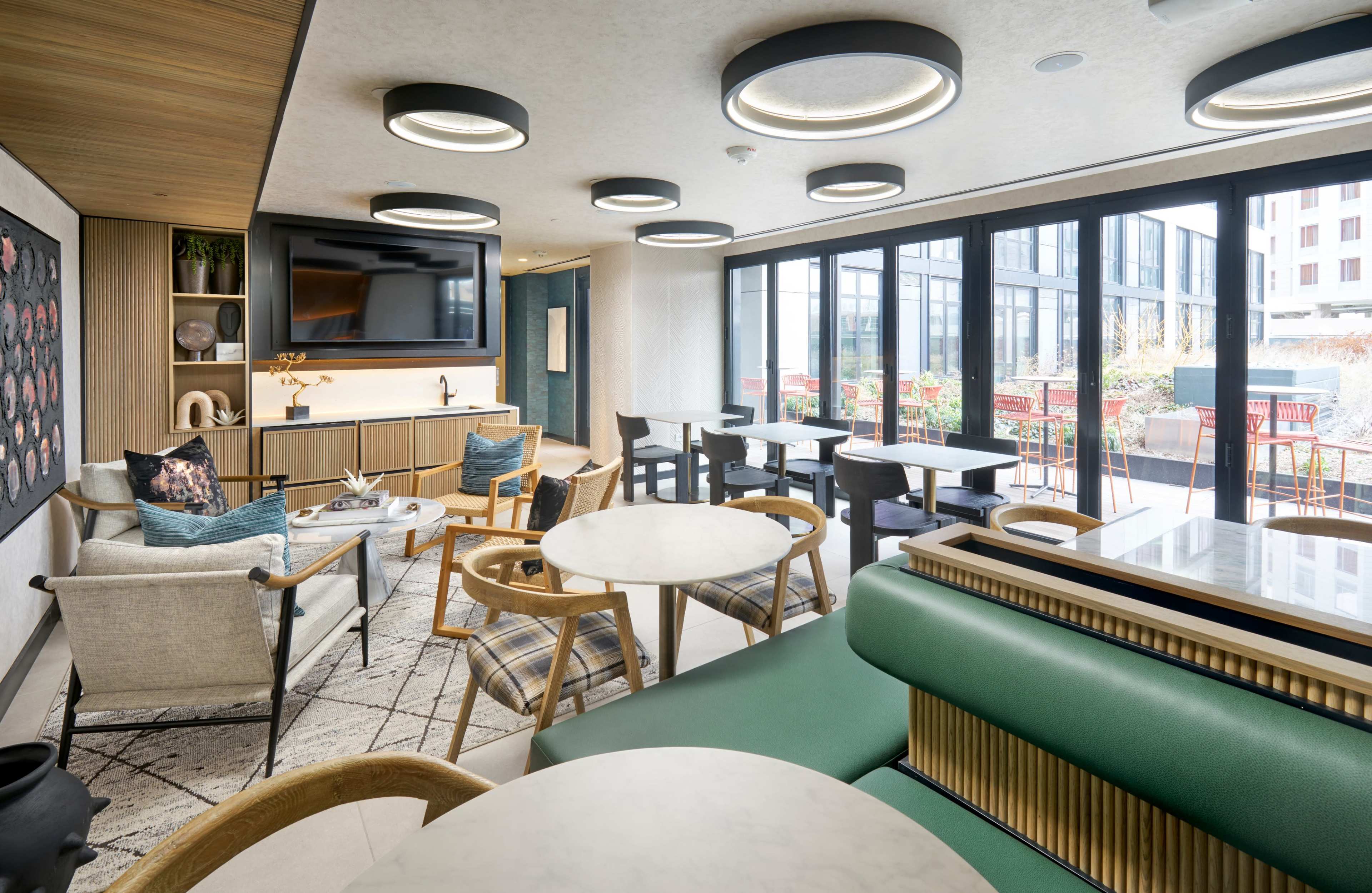 The image shows a modern lounge area with green seating, marble tables, and large windows overlooking an outdoor terrace.