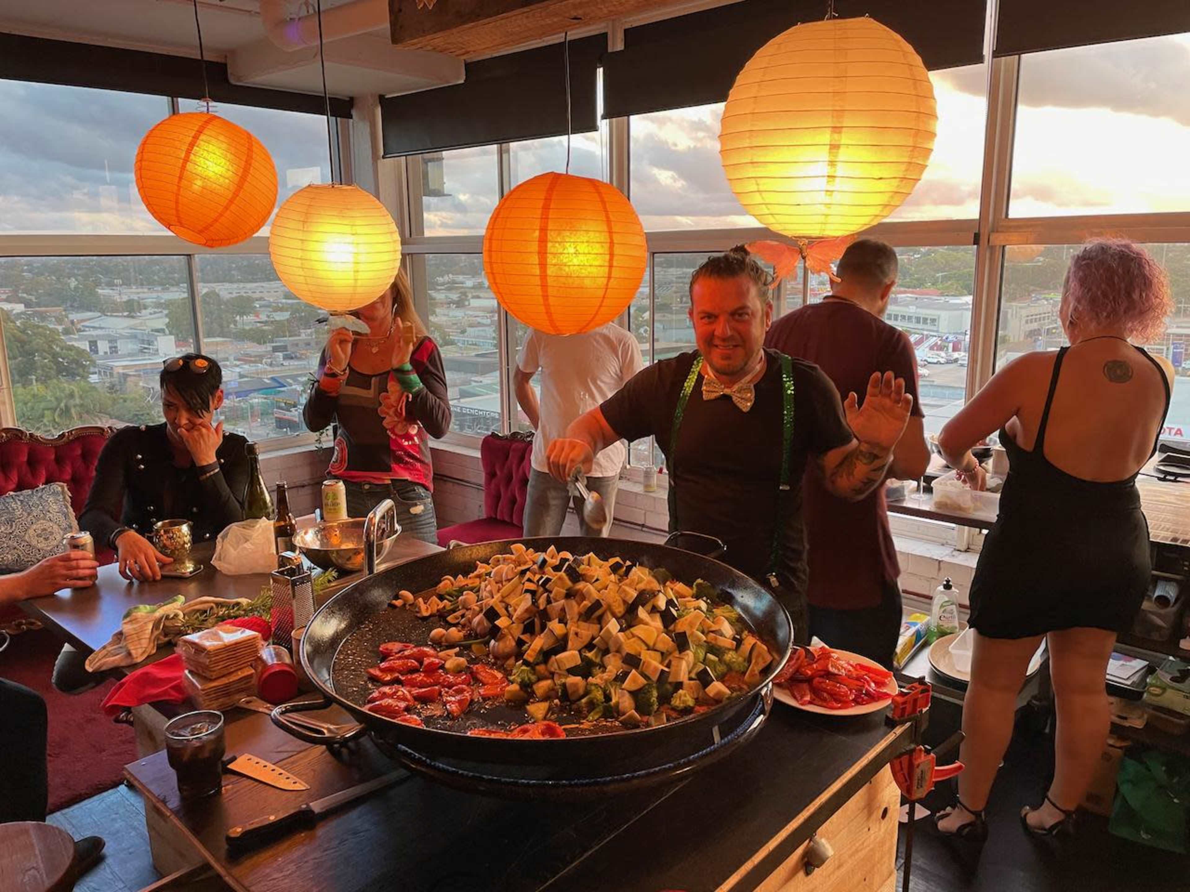 A man stands smiling next to a large pan of cooking food in a brightly lit room with hanging lanterns and people mingling around.