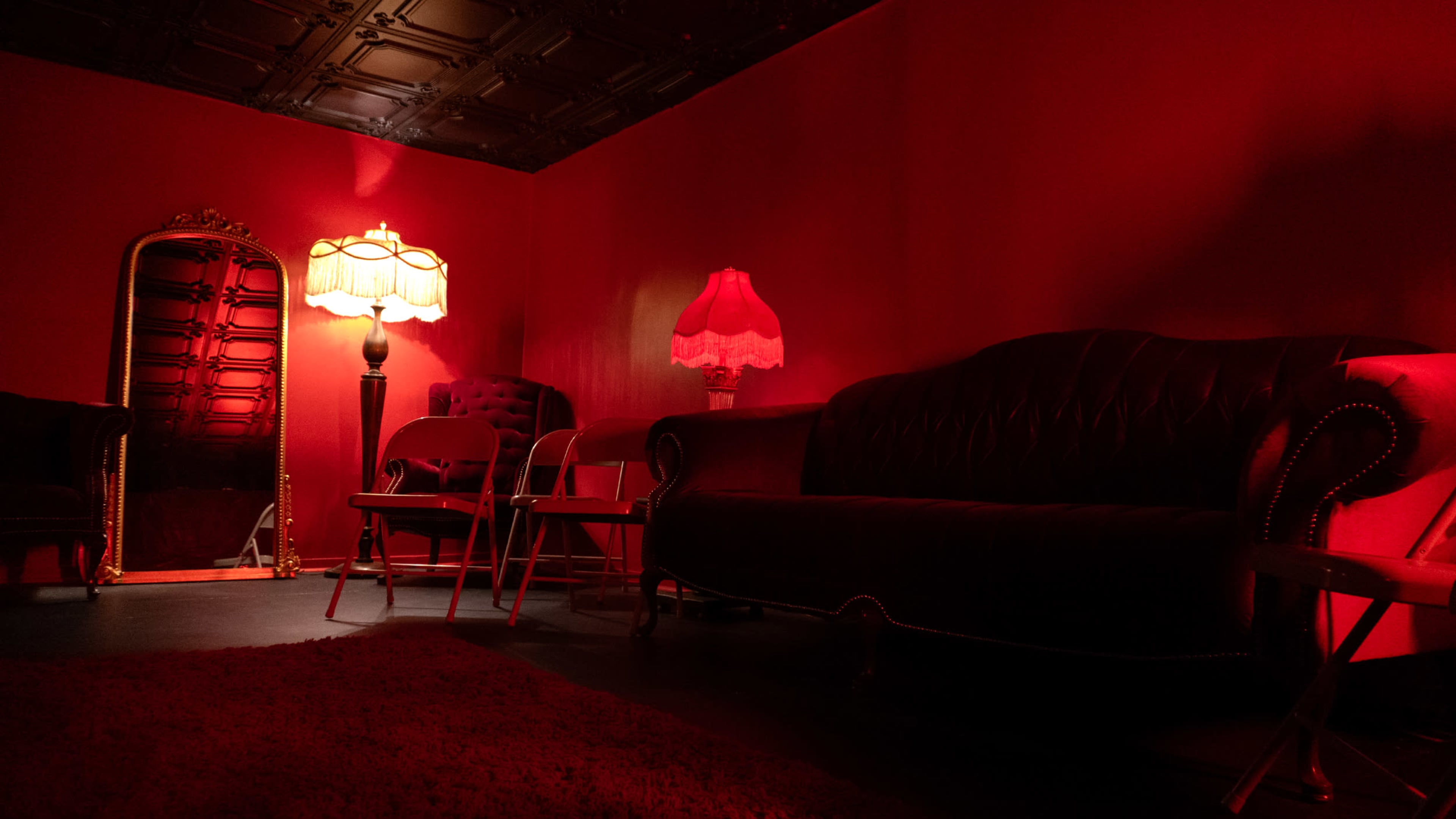 The scene features a red-lit room with a dark sofa, various chairs, and two lamps, one of which is a standing lamp and the other a table lamp.