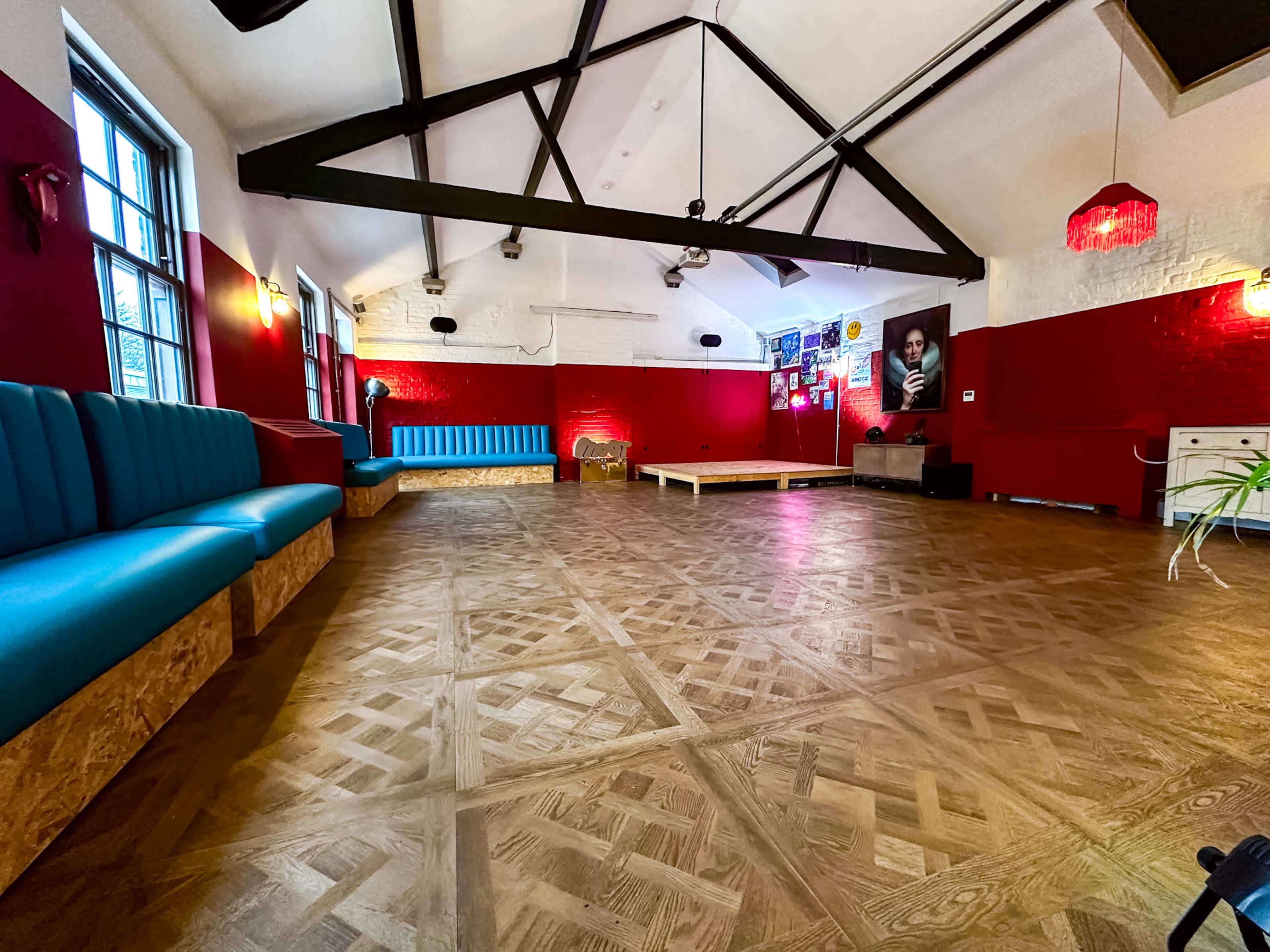 The image shows a spacious interior with wooden flooring, red and white walls, and various seating arrangements, including a blue couch and a wooden platform.