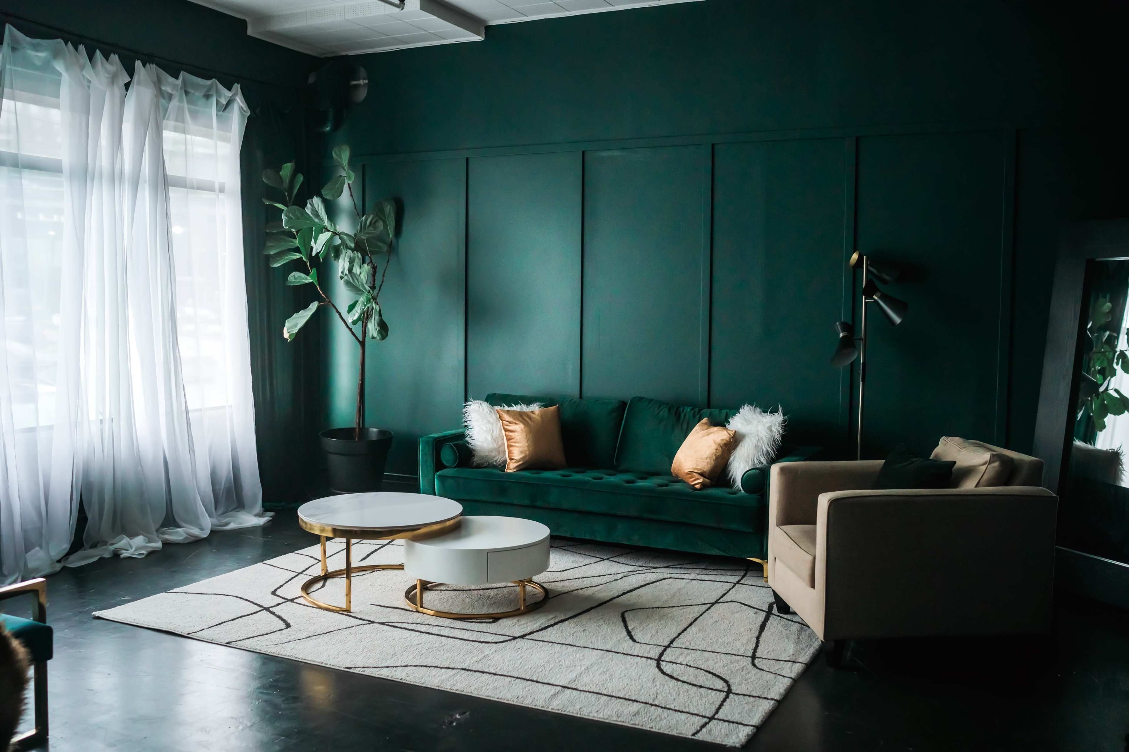 The image shows a modern living room with a dark green velvet sofa, a tan armchair, a round coffee table, and large windows with sheer white curtains.