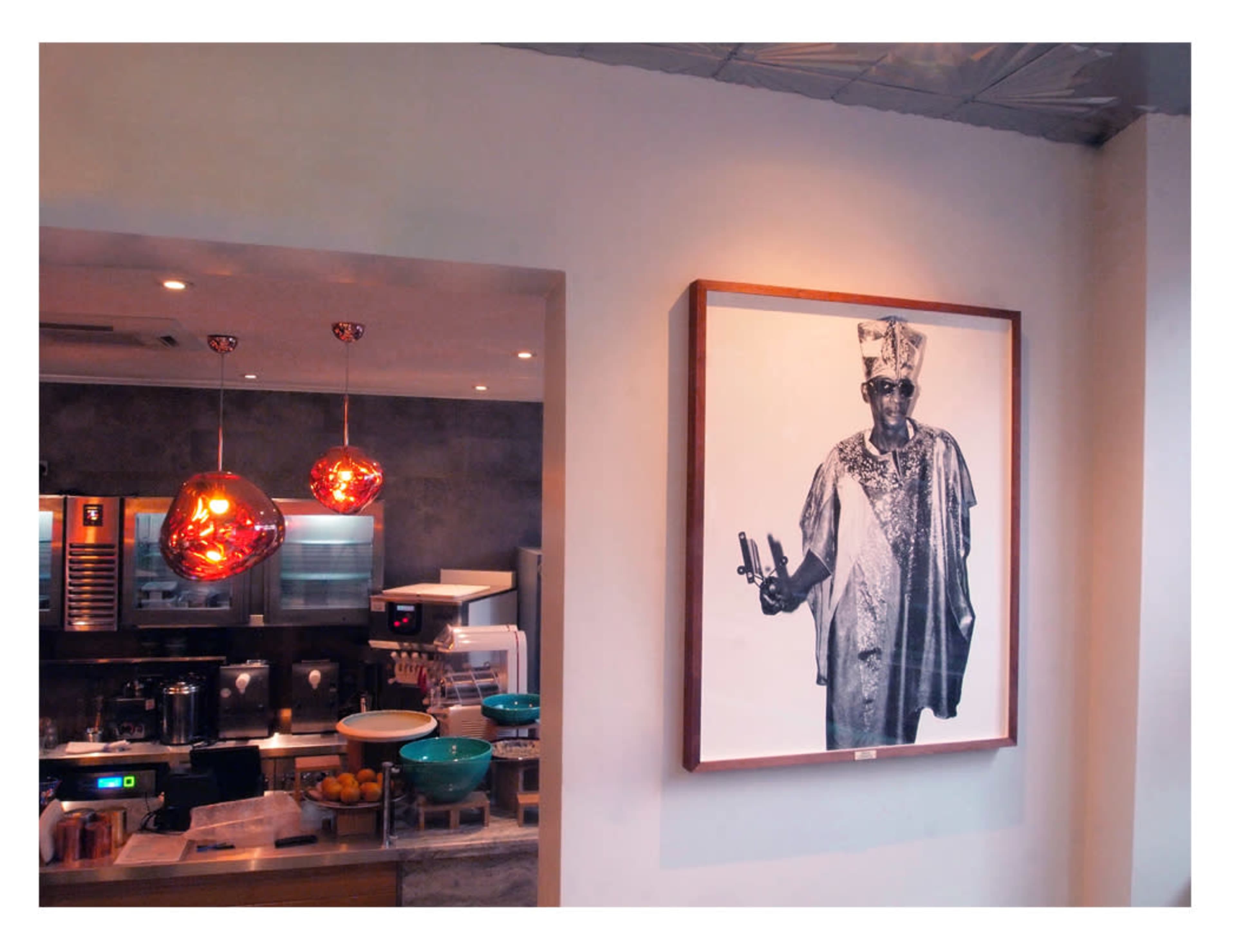 A restaurant interior with modern lighting fixtures, a kitchen area visible in the background, and a large black-and-white portrait of a man in traditional attire on the wall.