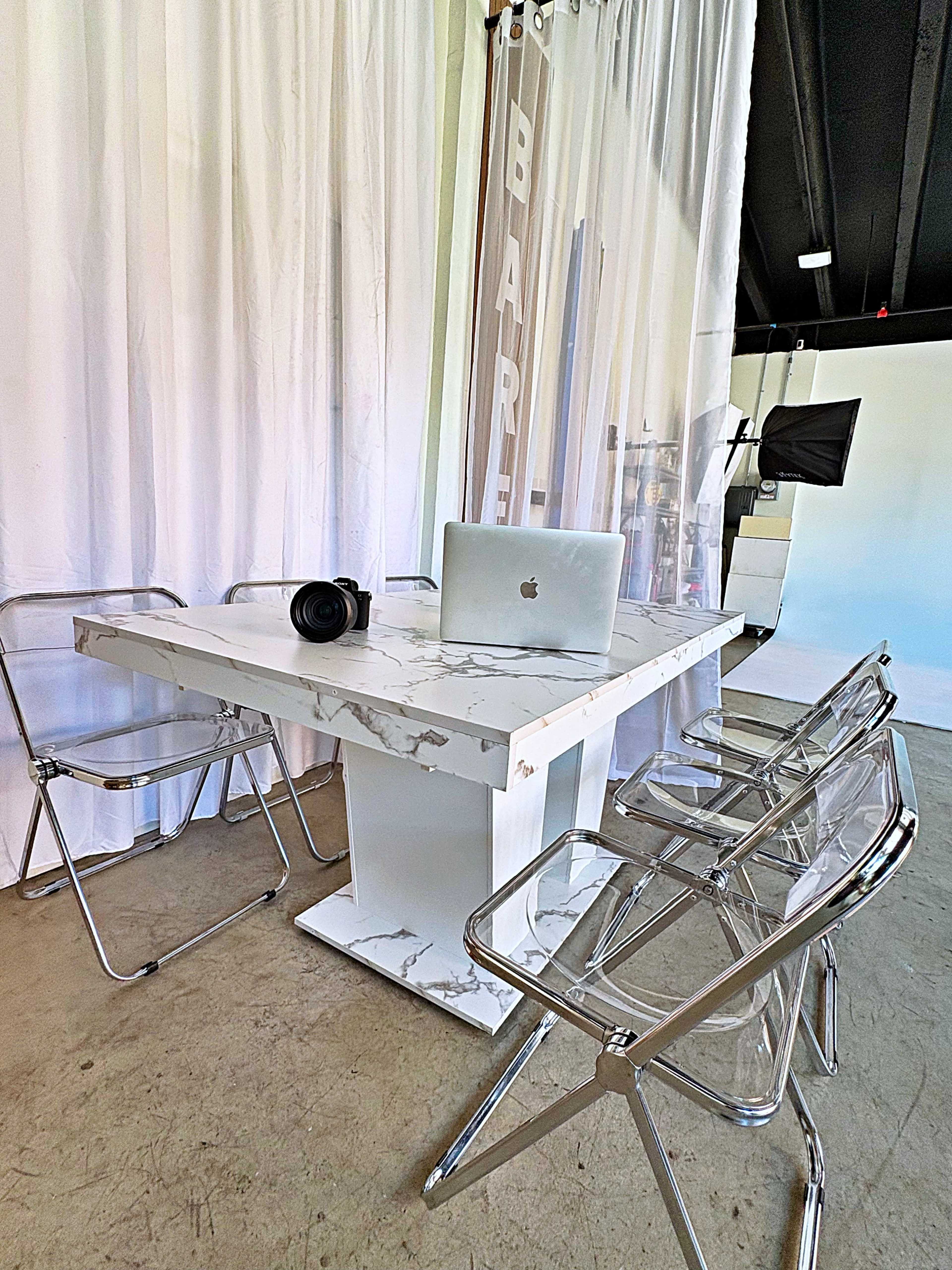 A modern workspace features a marble table surrounded by transparent chairs, with a laptop and camera placed on the table.