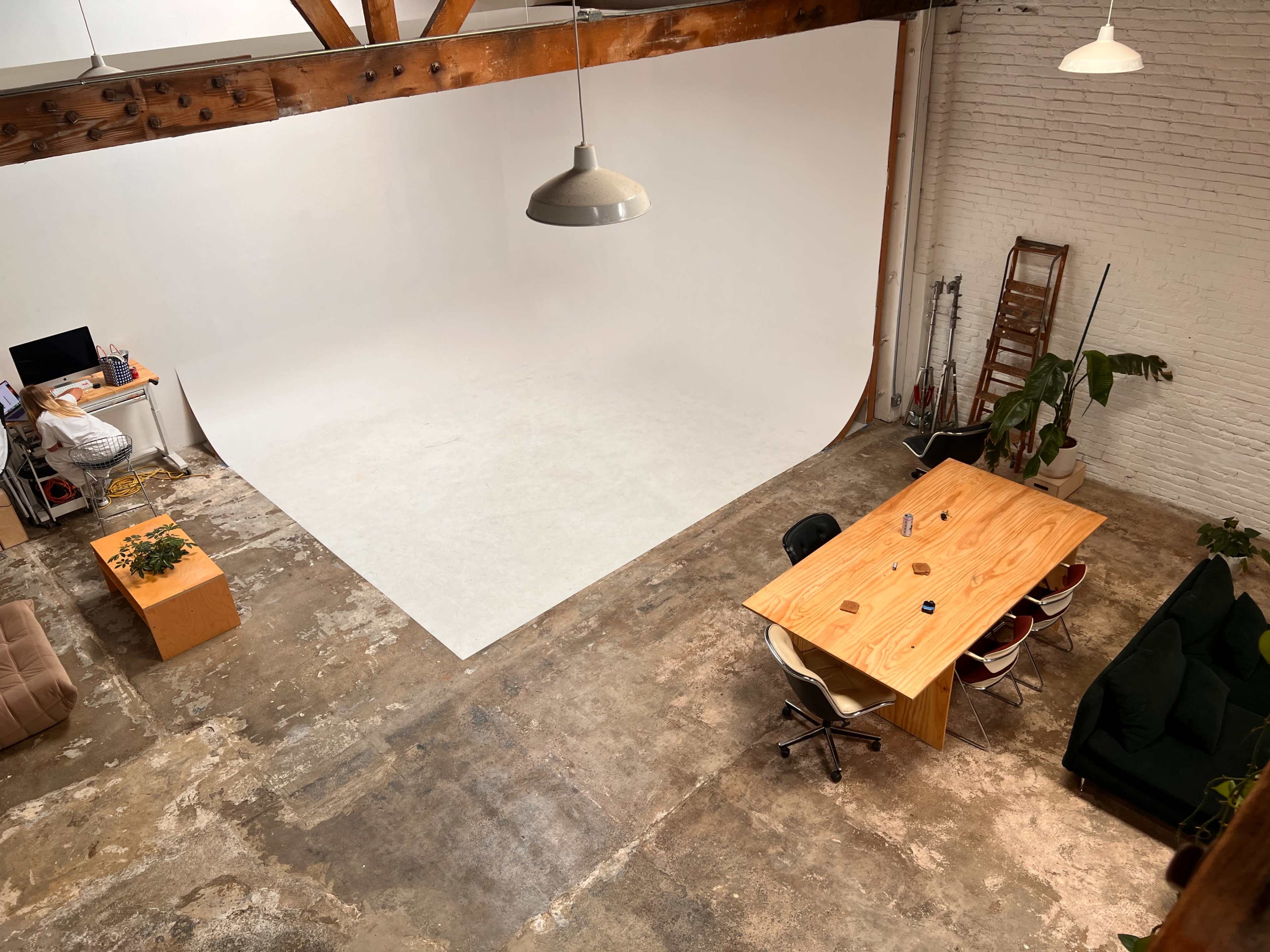 A spacious studio with a white backdrop, a wooden table surrounded by chairs, and plants along the walls.