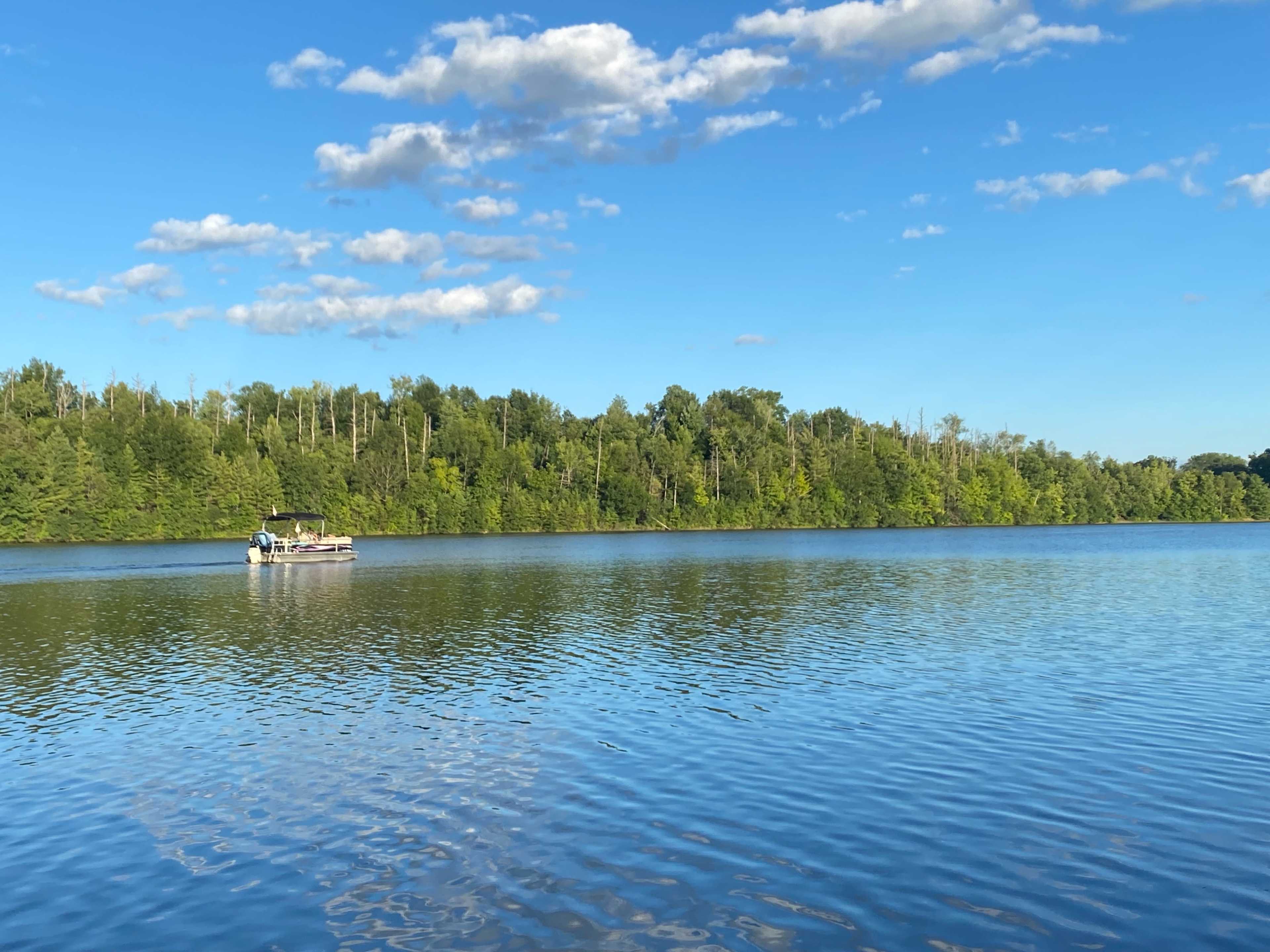 A boat is anchored on a calm lake surrounded by lush green trees under a blue sky with scattered clouds.