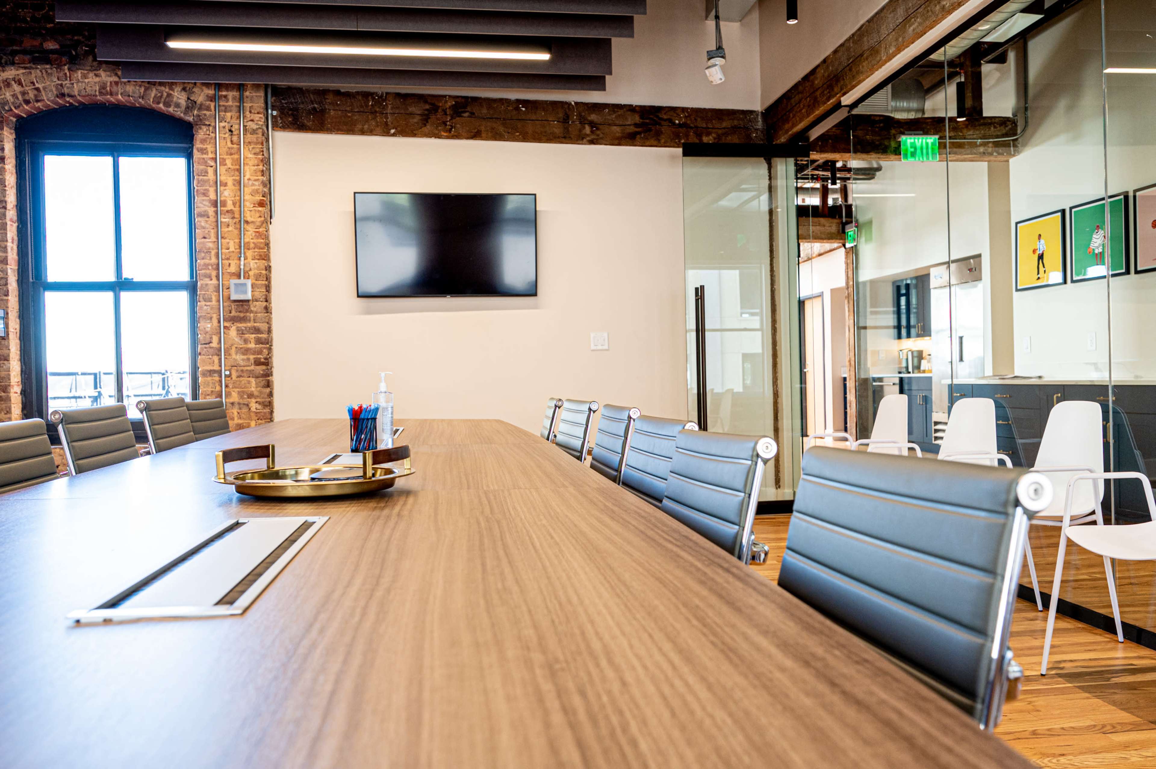 A modern conference room features a long wooden table, ergonomic chairs, a wall-mounted television, and large windows with exposed brick.