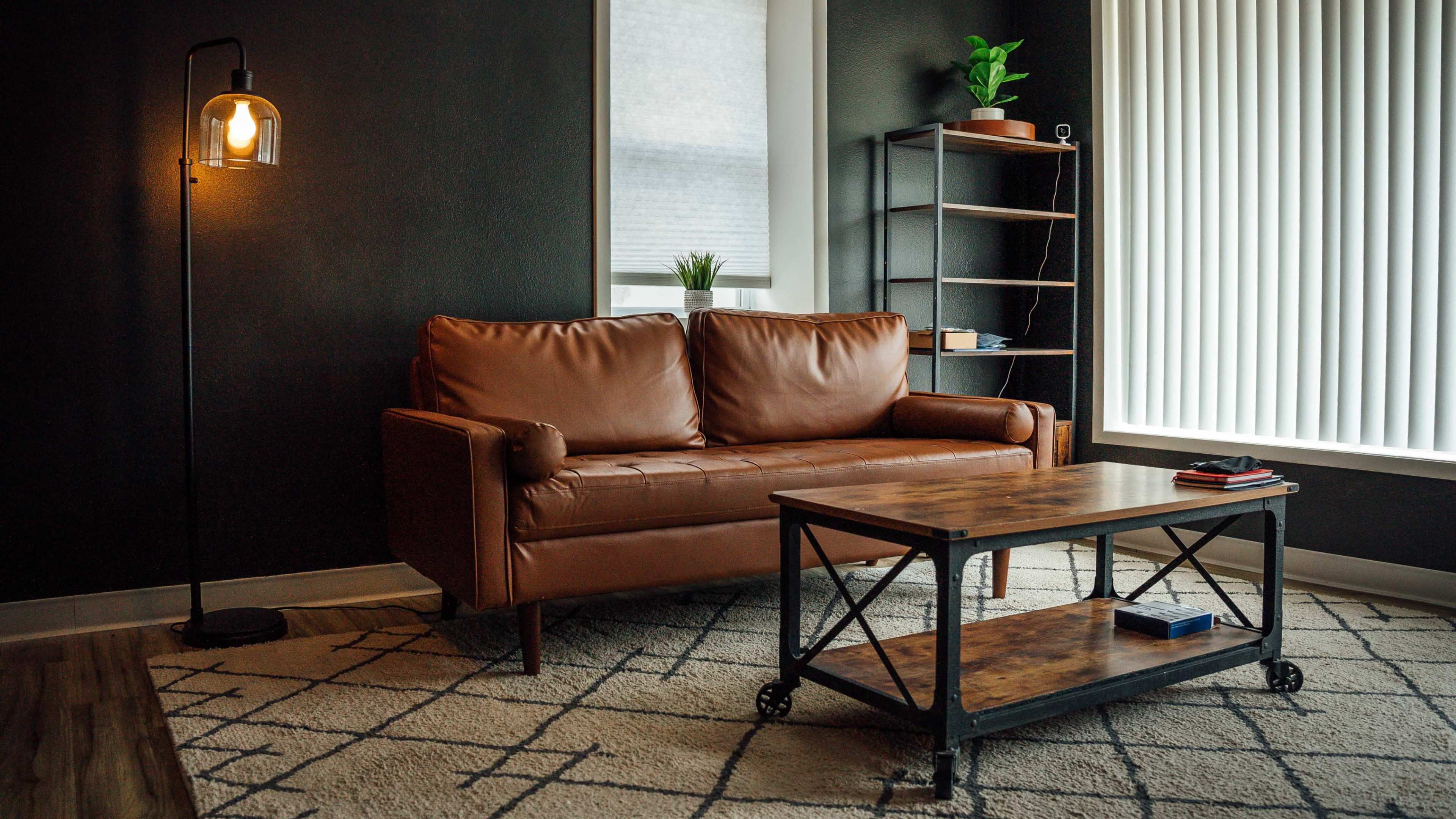 A brown leather sofa is positioned across from a wooden coffee table on a geometric-patterned rug in a room with dark walls and a tall, slim bookshelf.