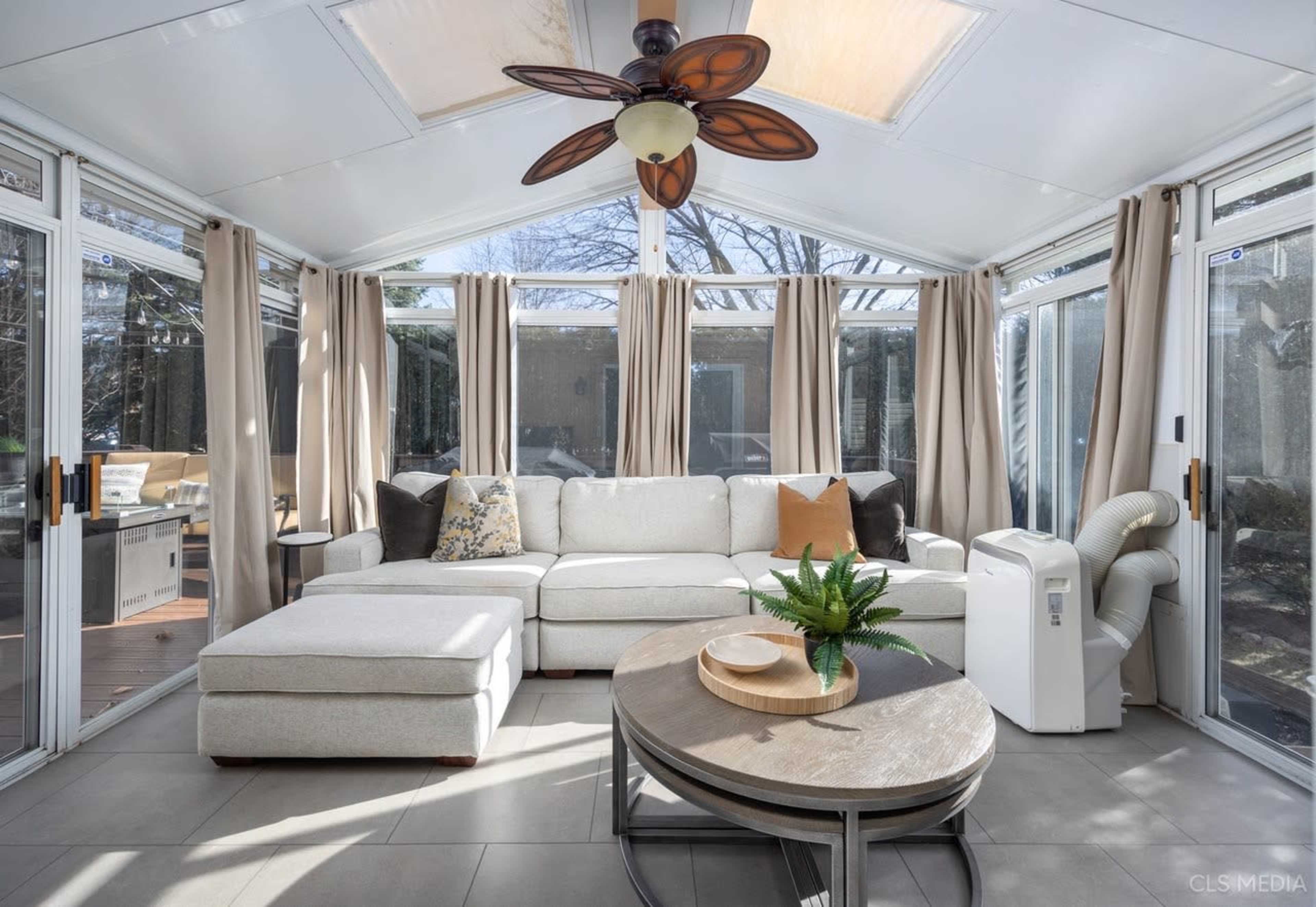 The image shows a sunroom featuring a large sectional sofa, a round coffee table with a plant, and large windows with beige curtains.