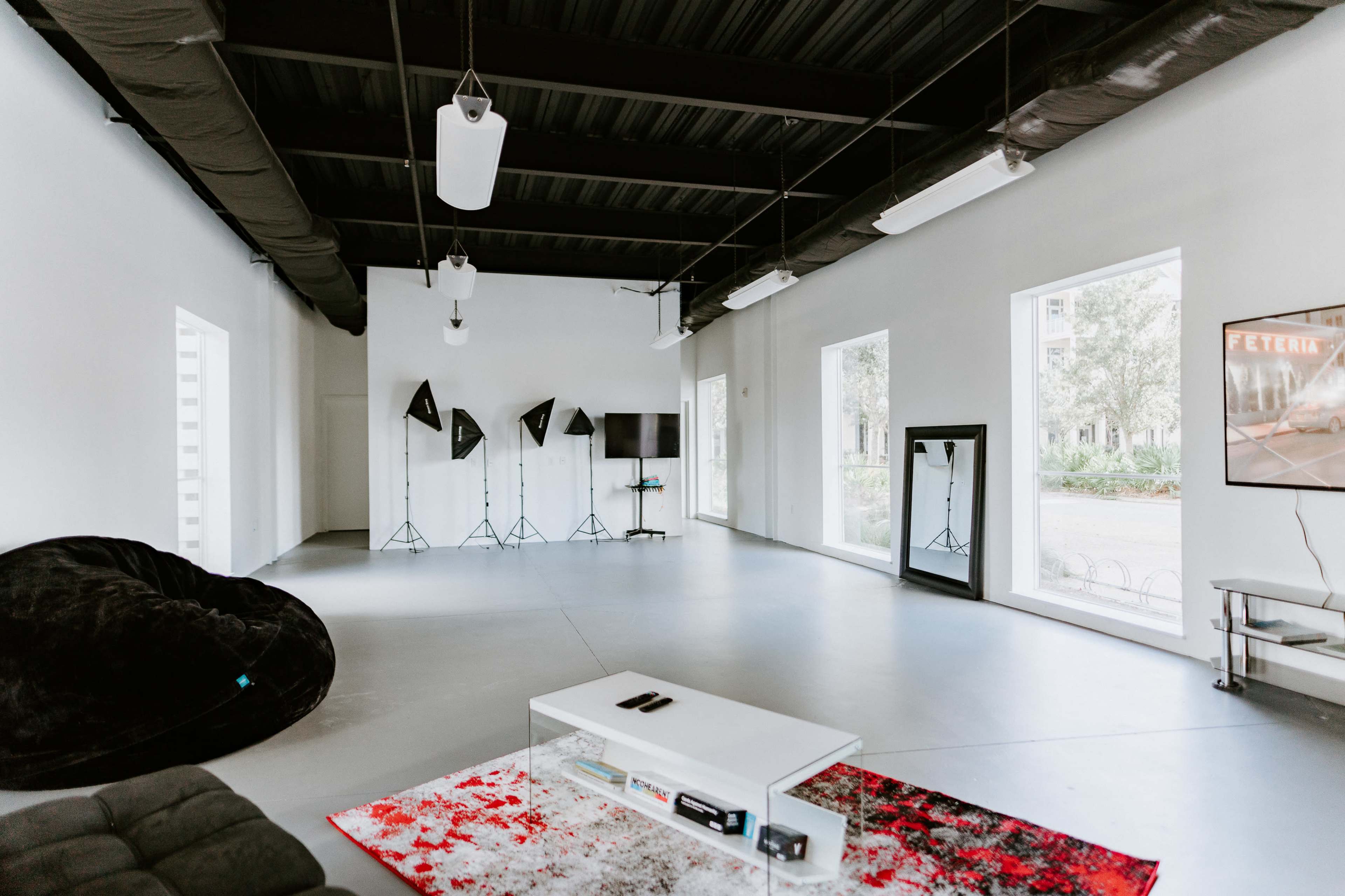 A spacious, well-lit studio with a large black bean bag, white walls, a coffee table, and several photography lights positioned around the room.