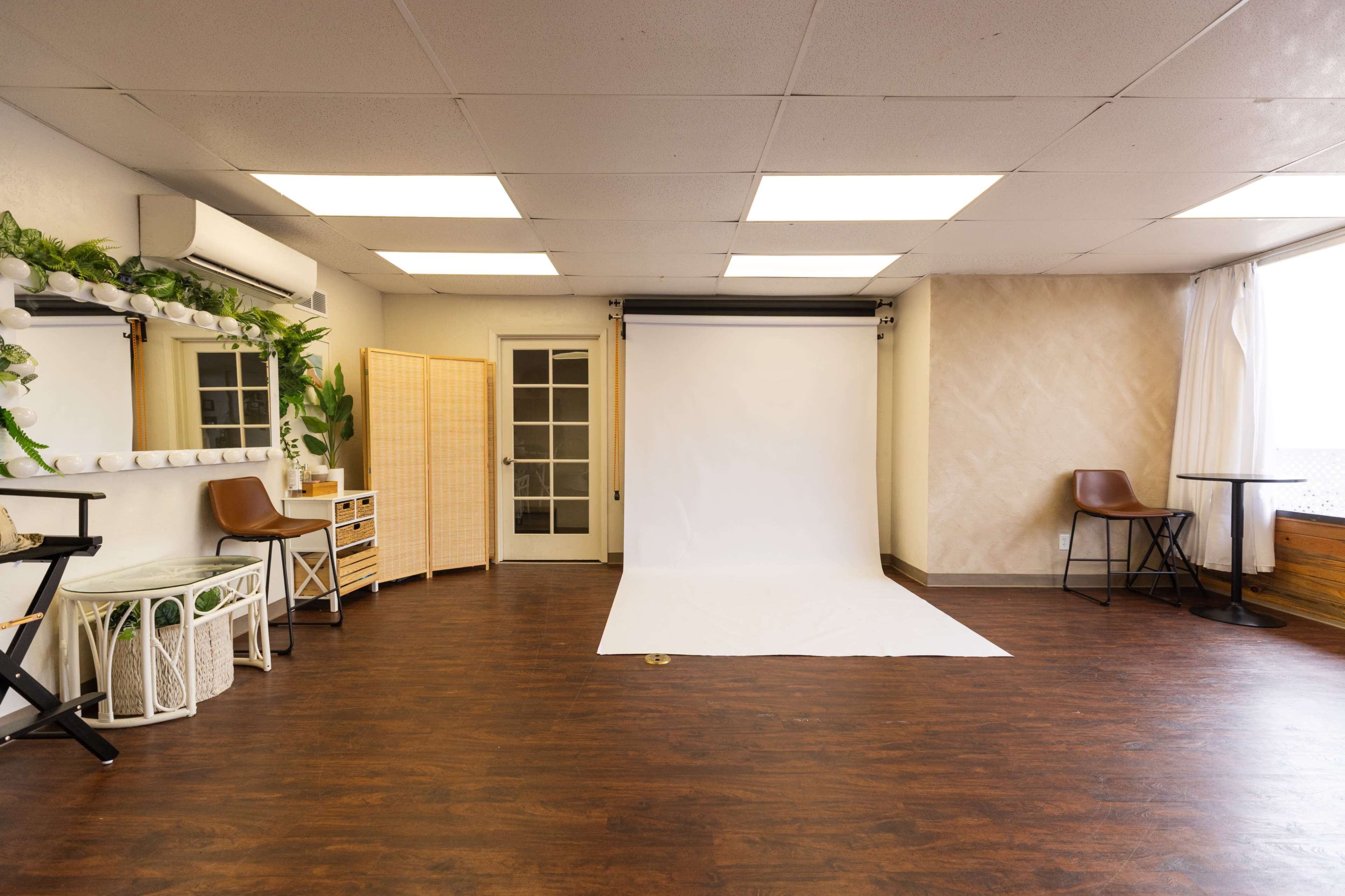 The image shows a spacious studio with a white backdrop, wooden flooring, and minimalist furniture, including chairs and a storage cabinet.