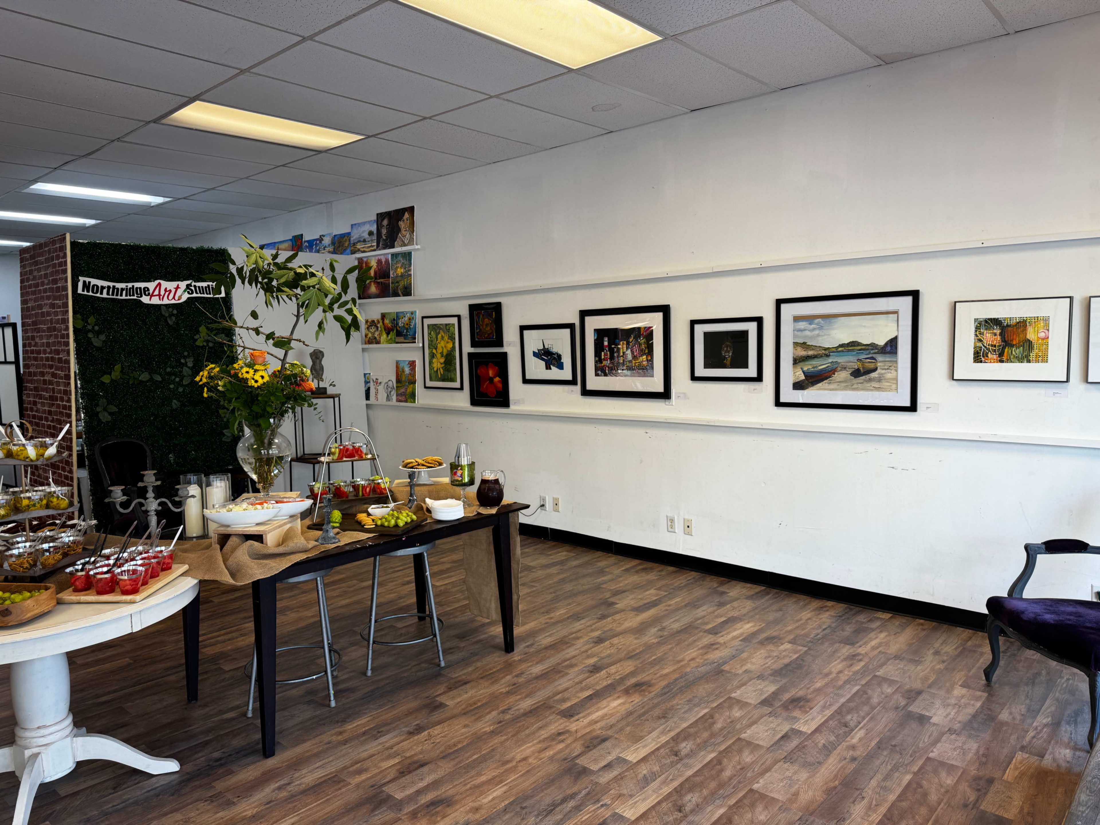 The image shows an art gallery interior featuring a display of framed artworks on the wall, a table set with food, and a decorative arrangement of flowers.