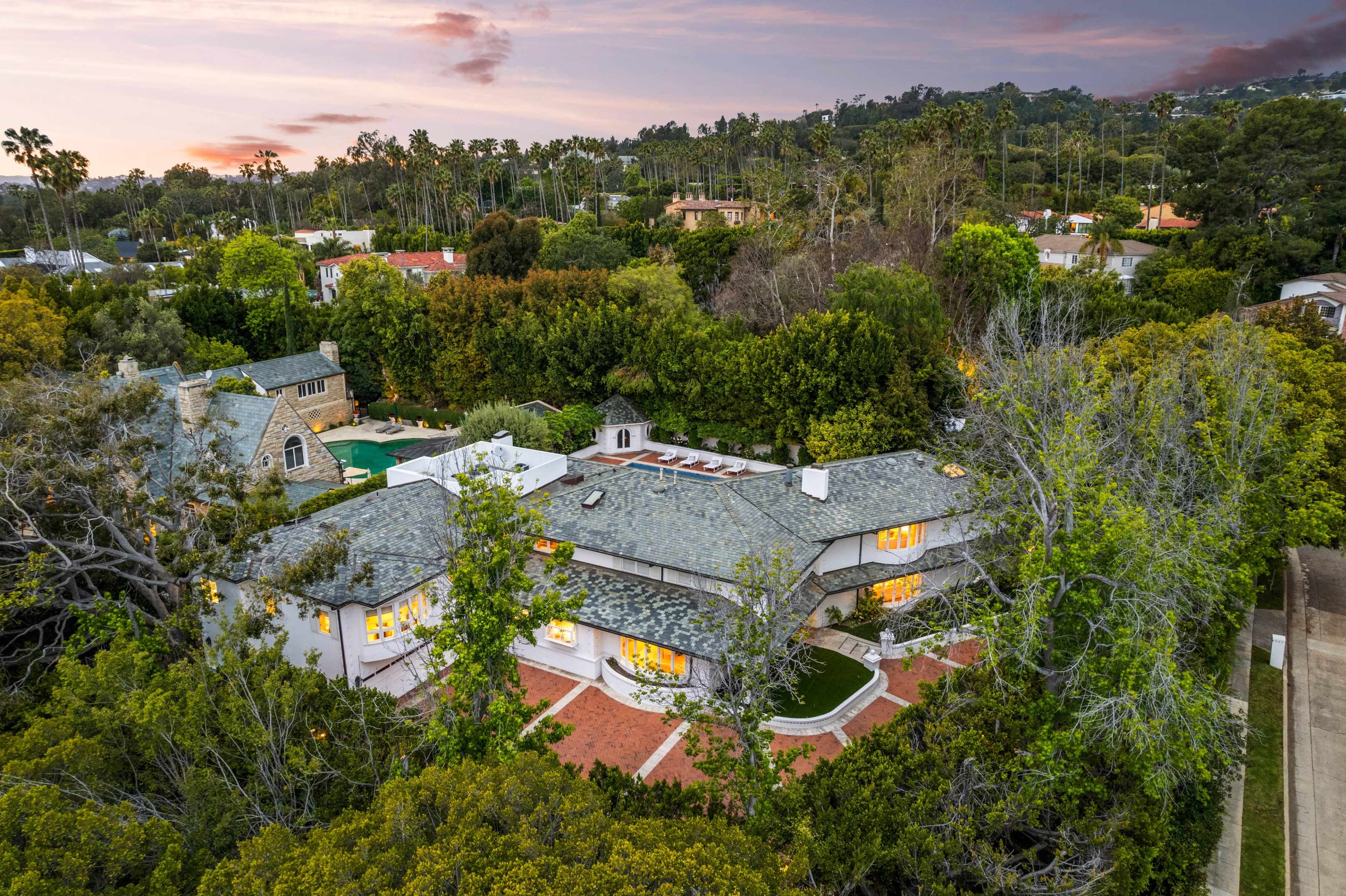 A spacious house with a slate roof is surrounded by lush greenery and trees, set against a backdrop of a sunset sky.