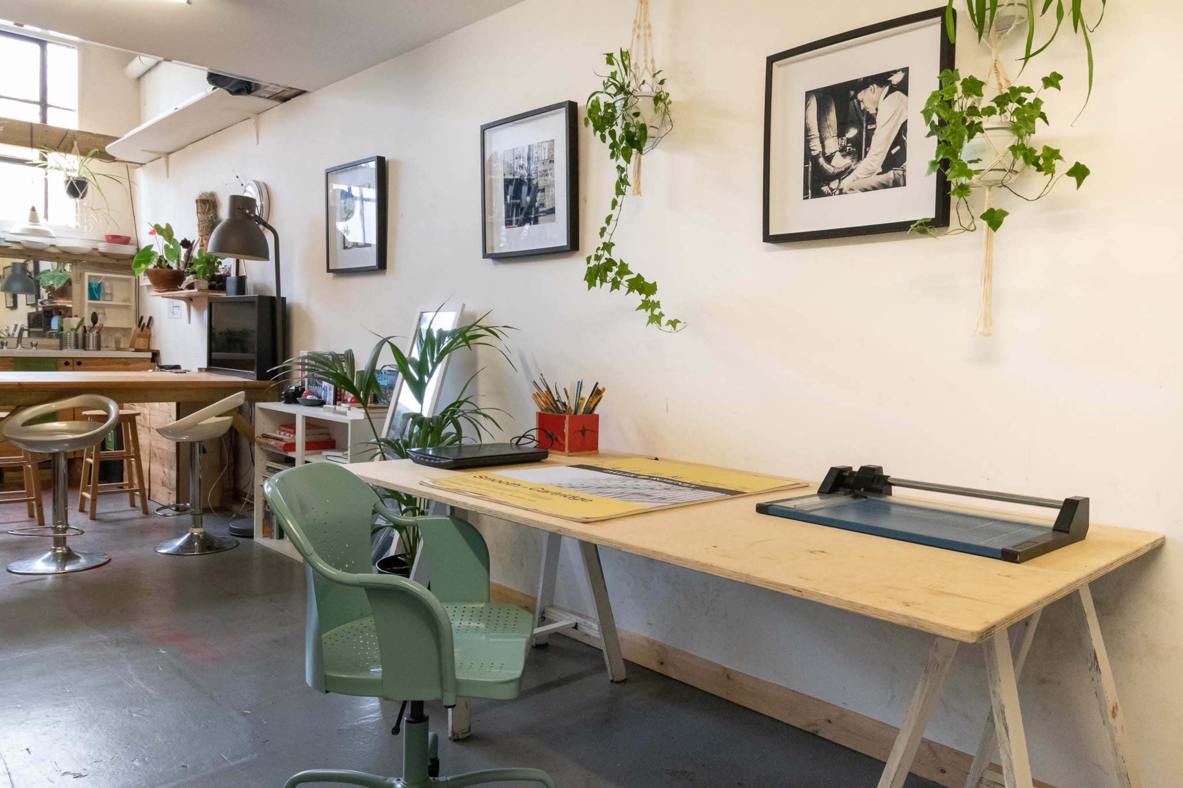 A workspace with a wooden table, a green chair, framed photographs on the wall, and plants hanging from the ceiling.