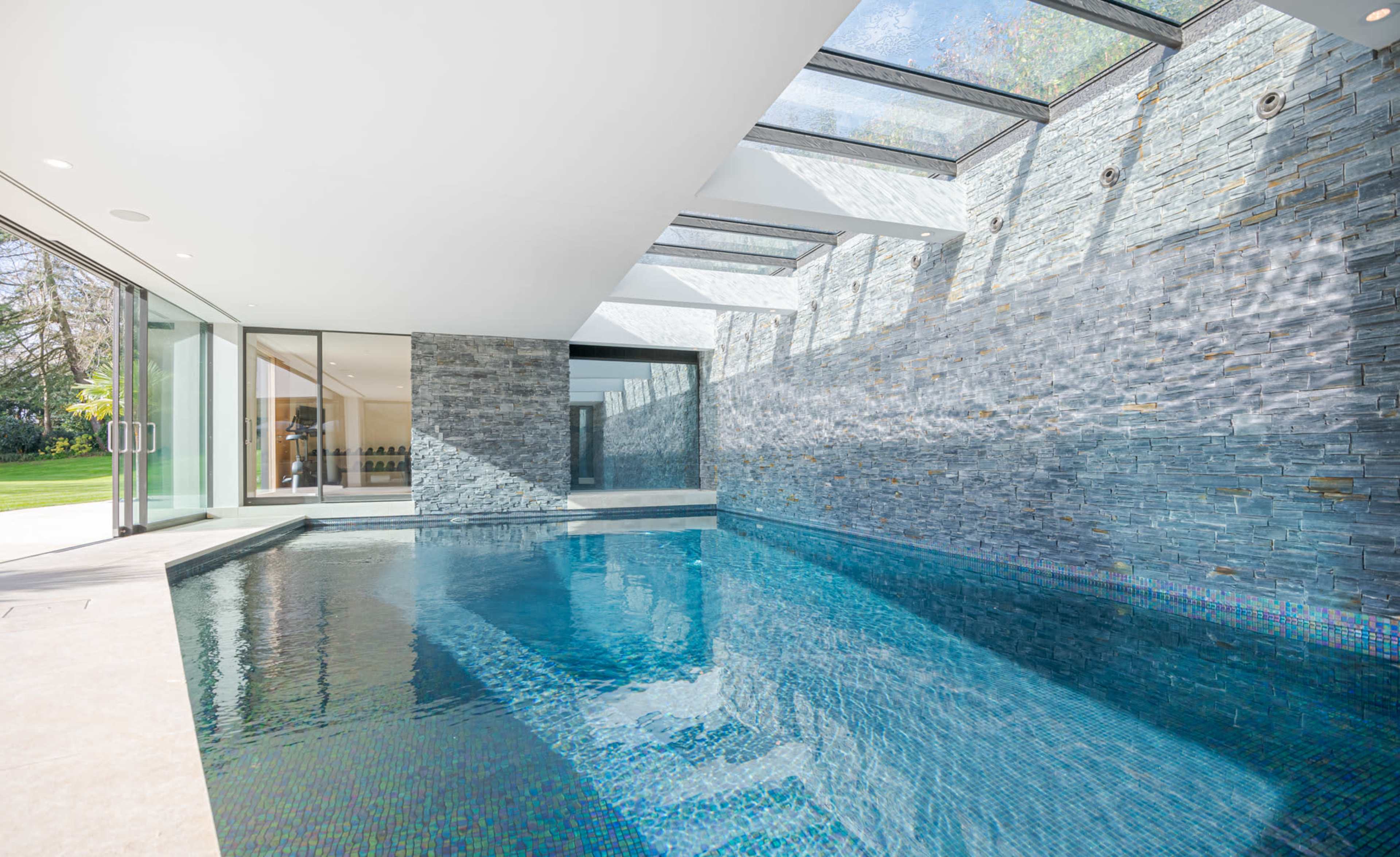 The image shows an indoor swimming pool surrounded by walls made of stone and large glass panels letting in natural light.